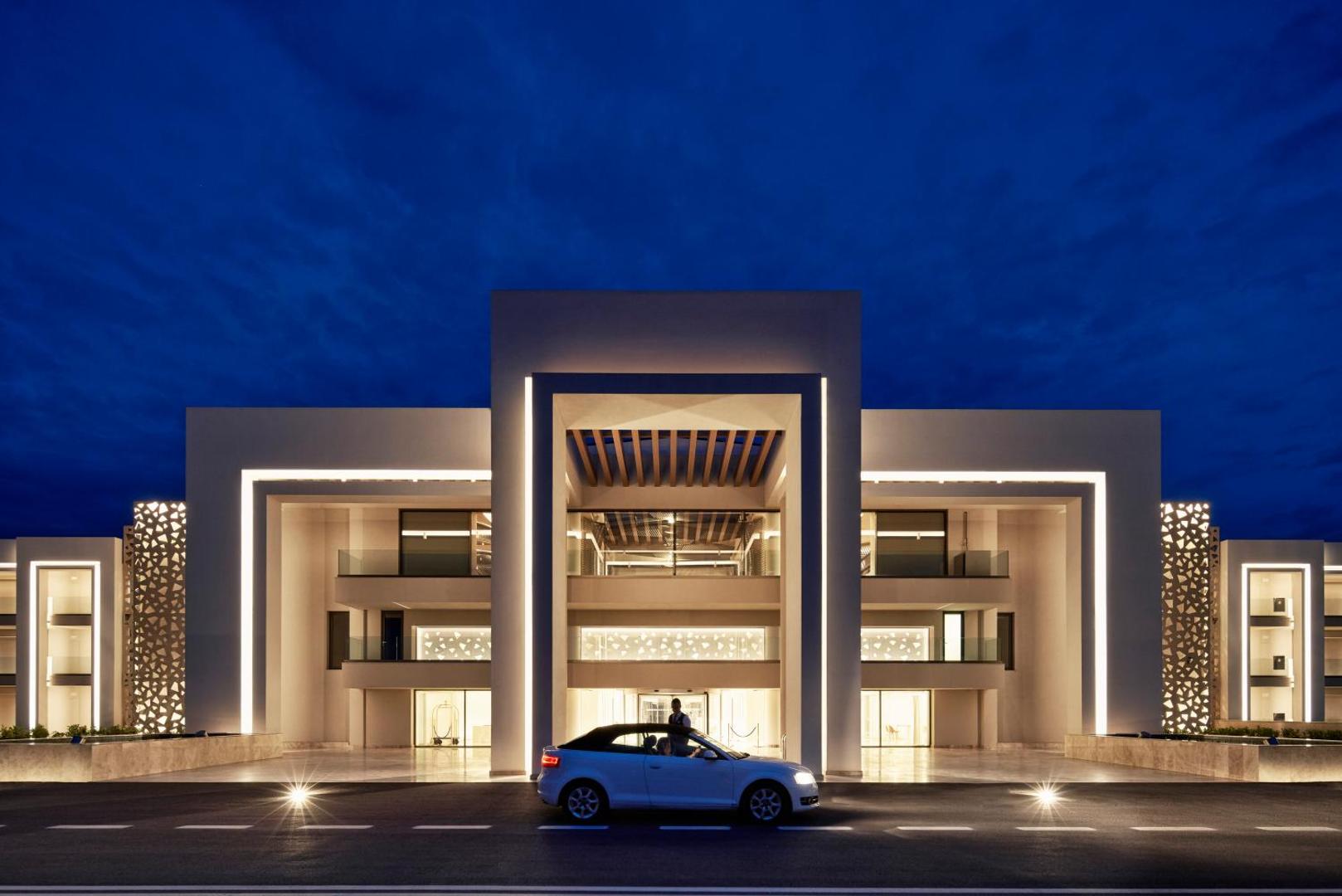 Mayia Exclusive Resort & SPA galvenā ieeja vakarā ar modernu apgaismojumu un baltu automašīnu priekšplānā Rodas salā