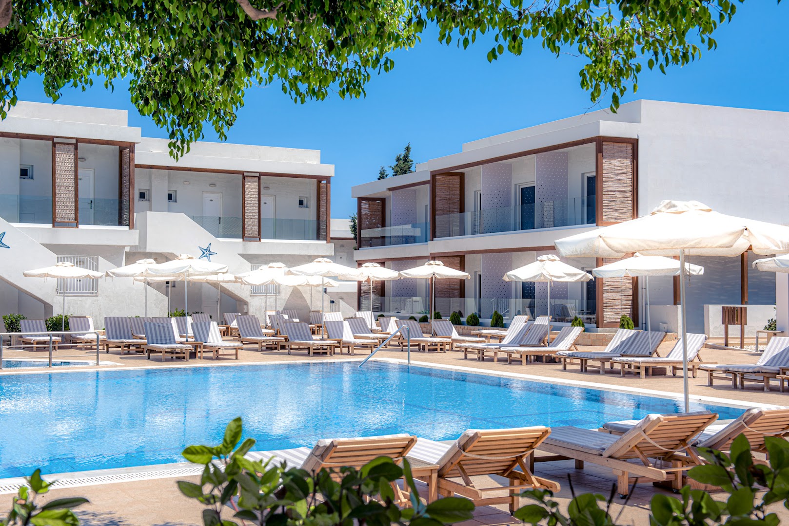 Aelius Hotel and Spa āra baseins ar atpūtas krēsliem un saulessargiem pie baltām divstāvu ēkām Grieķijā, Krētas salā.
