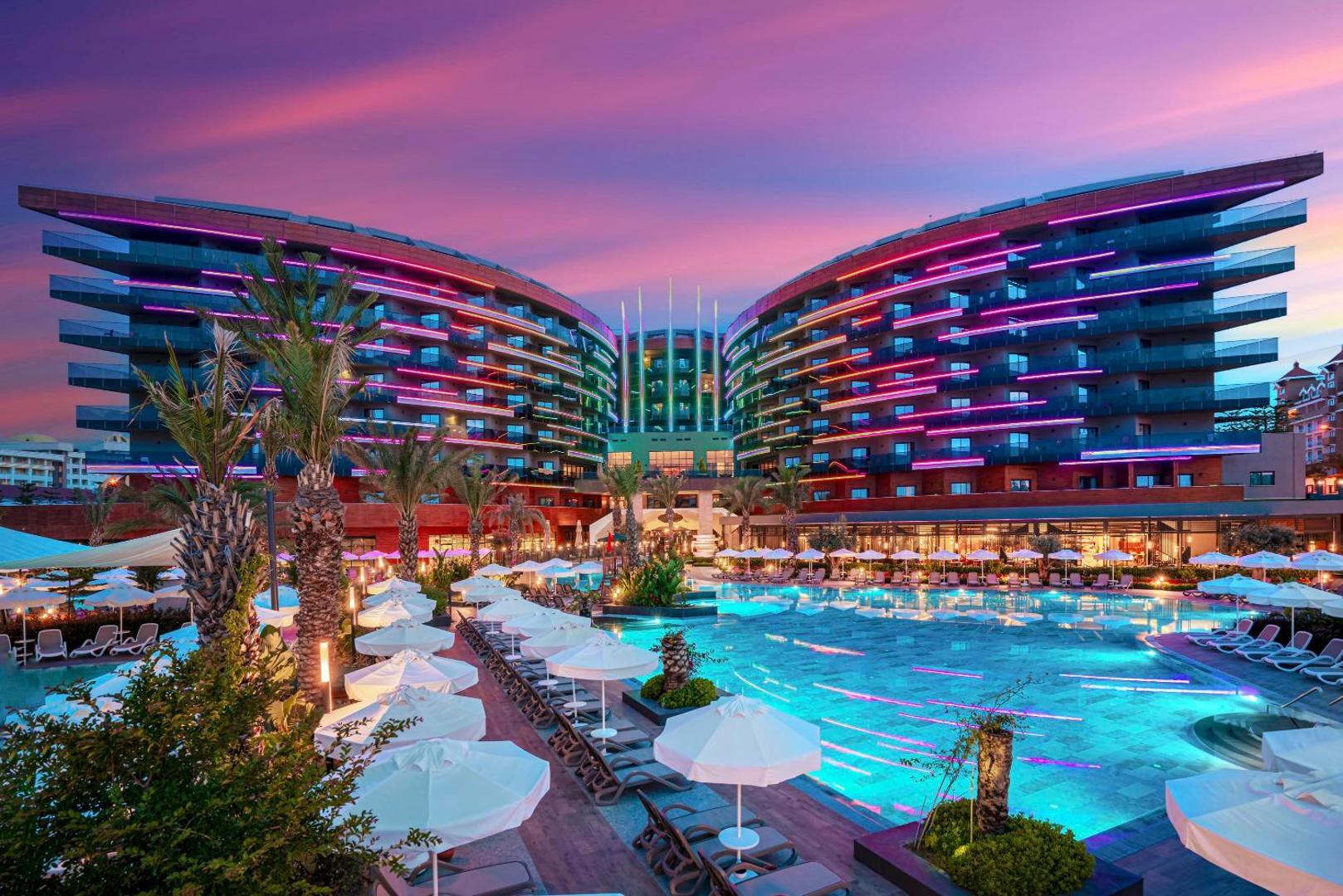 Kirman Calyptus Resort & SPA viesnīcas ēka un galvenais baseins vakarā ar krāsainu neonu apgaismojumu un saulrieta debesīm