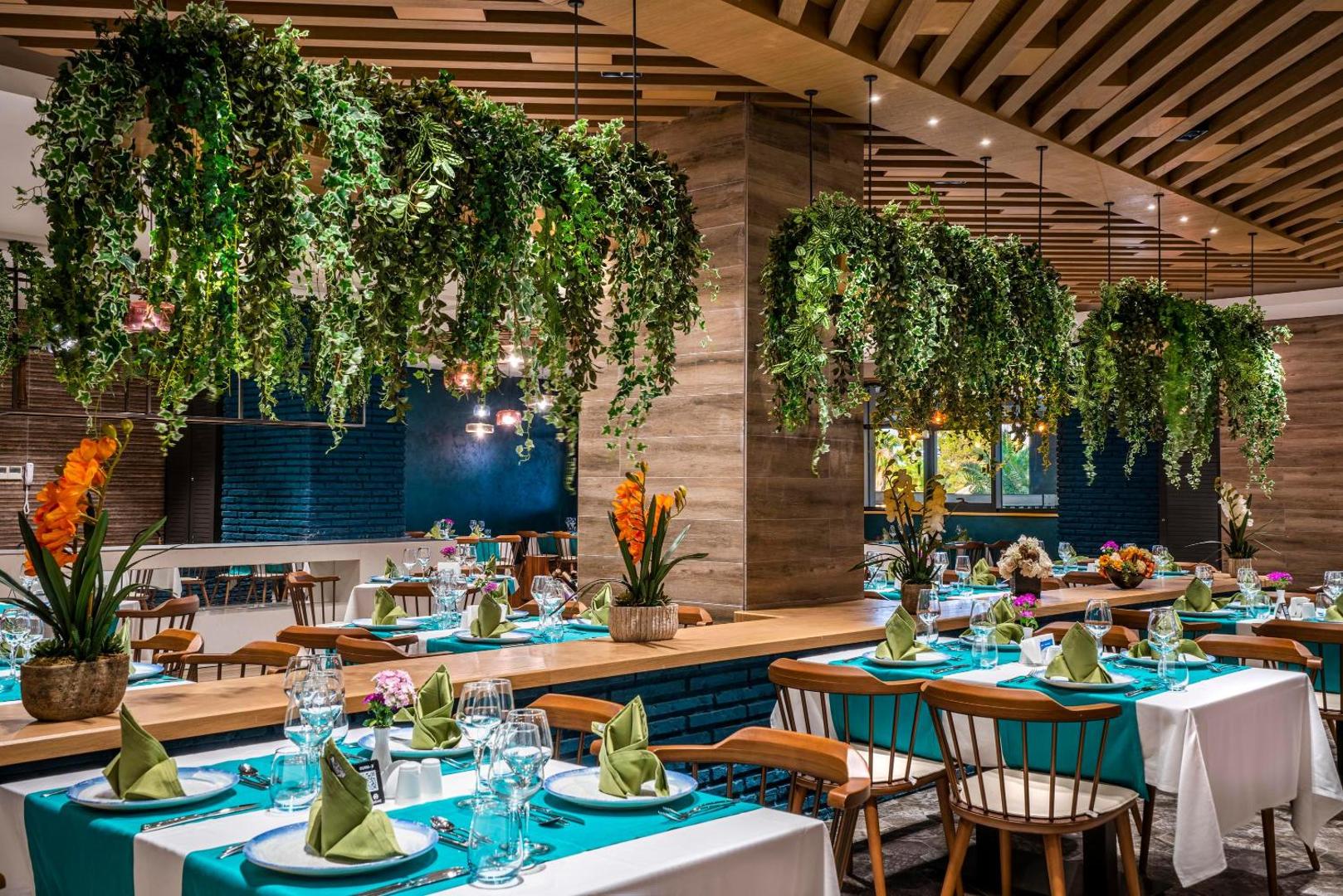 Kirman Calyptus Resort & SPA restorāns ar modernu dizainu, nokarenajiem telpaugiem, koka elementiem un eleganti klātiem galdiem tirkīza toņos
