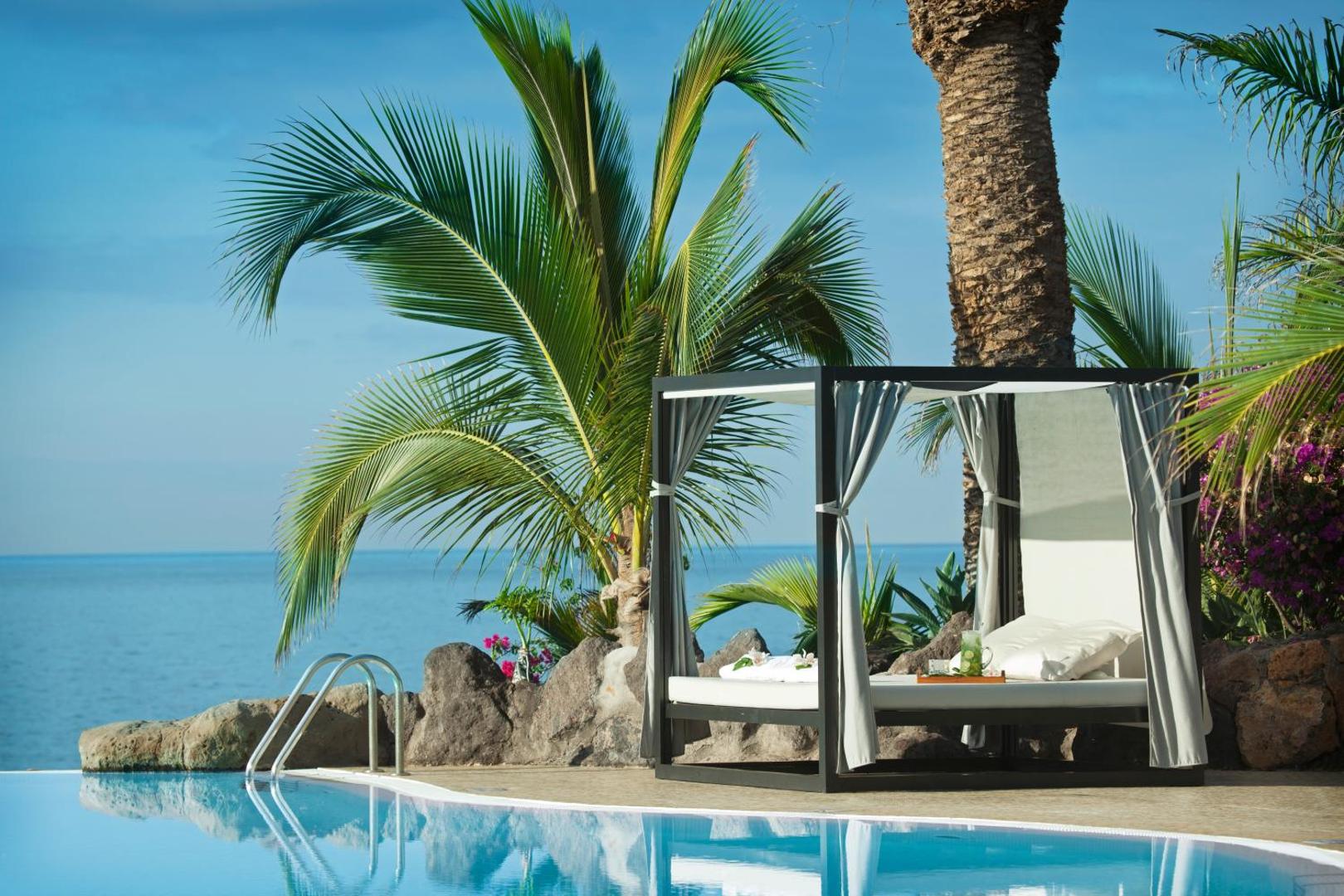 Ekskluzīva atpūtas gulta pie Roca Nivaria Gran Hotel baseina Tenerifē, ieskauta palmās ar skatu uz zilo jūru