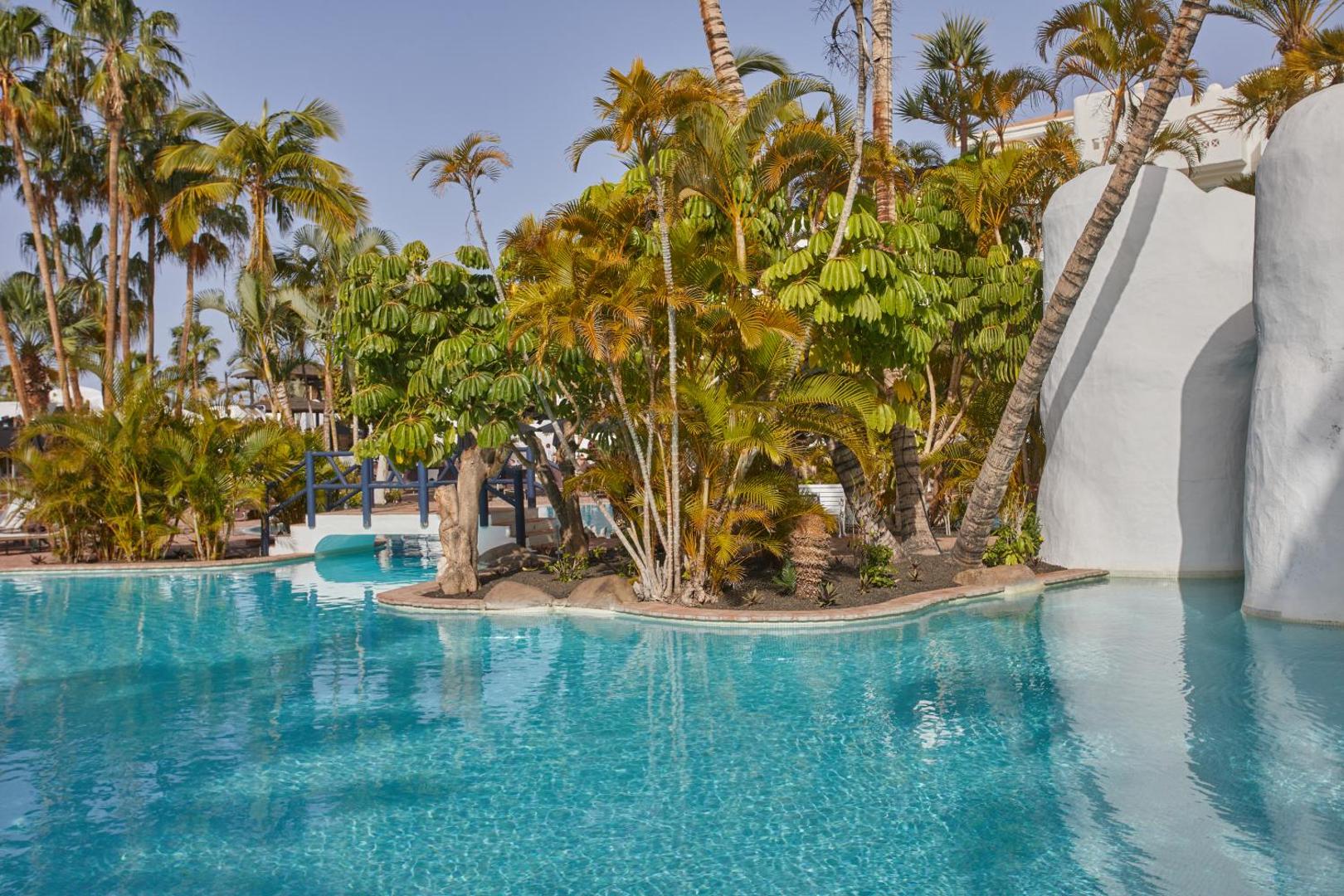 Dreams Jardin Tropical Resort & Spa plašais āra baseins ar tirkīza ūdeni, ko ieskauj eksotiski augi, palmas un balti arhitektūras elementi