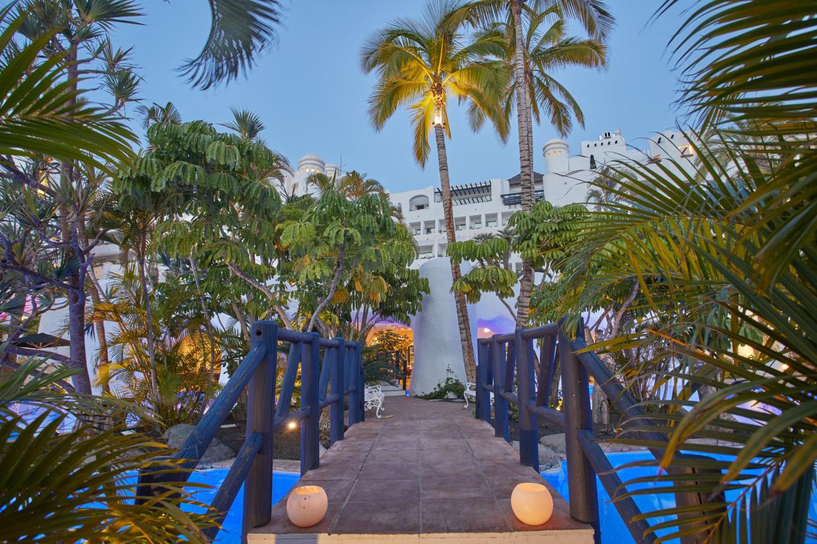 Dreams Jardin Tropical Resort & Spa vakara apgaismojums dārzā ar izgaismotu tiltiņu, baseinu un palmām Tenerifes piekrastē