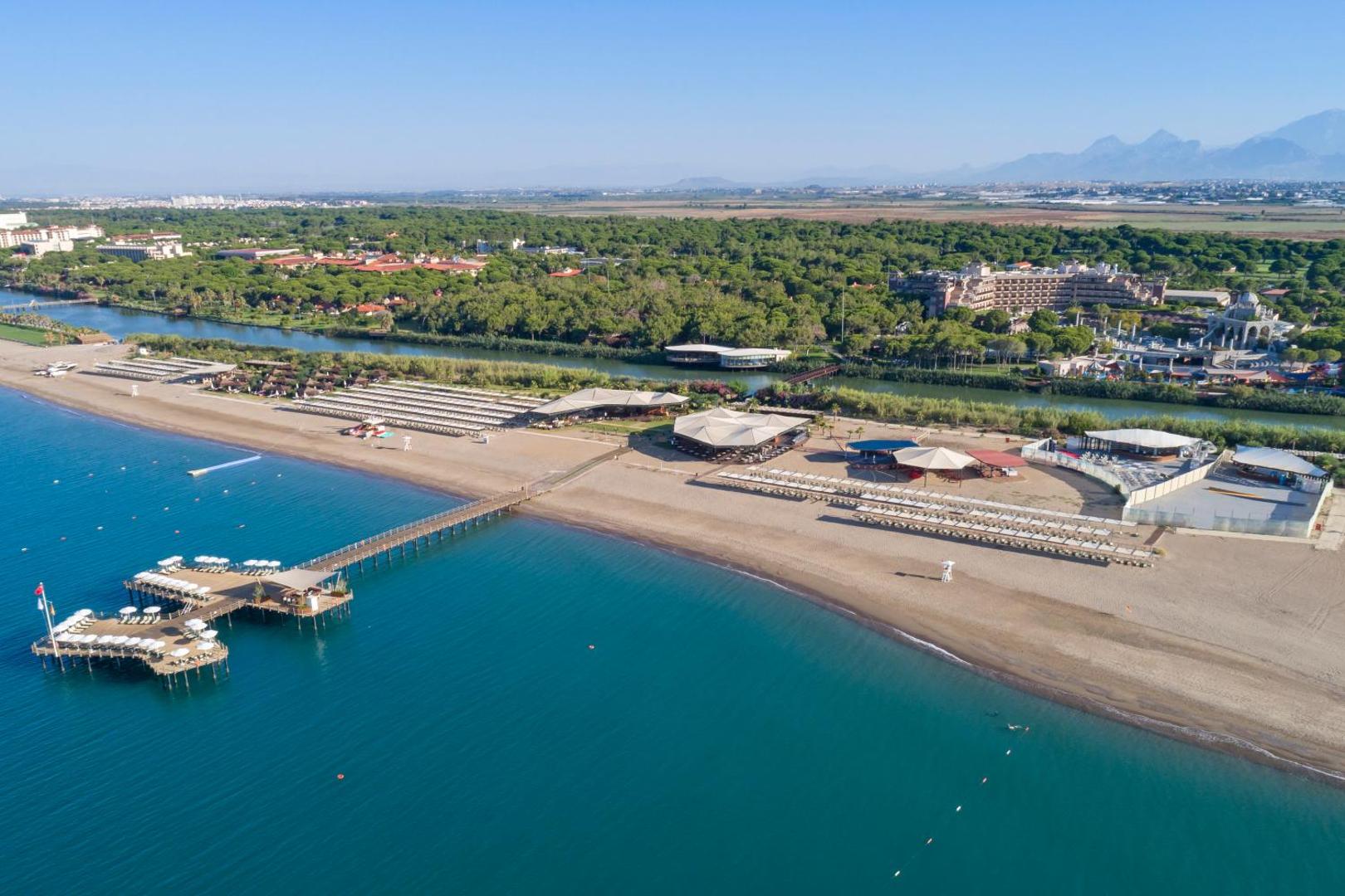 Xanadu Resort Hotel aerofotoattēls Belekā, Turcijā — plaša smilšu pludmale, koka piestātne un viesnīcas ēka priežu meža ielokā