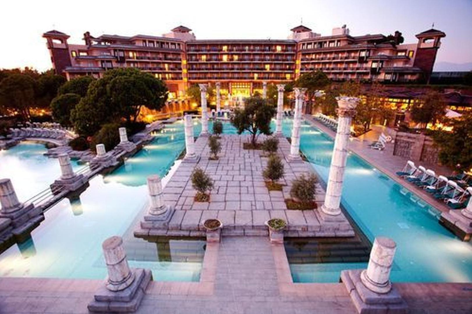 Xanadu Resort Hotel galvenā ēka un plašais baseinu komplekss ar romiešu stila kolonnām saulrietā Belekas reģionā