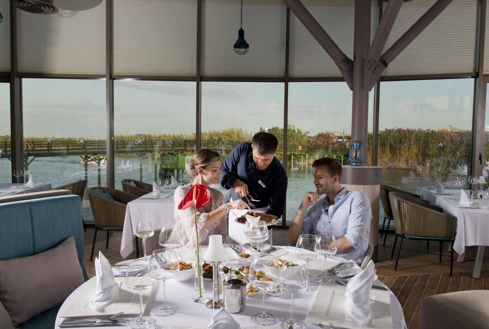 Viesi bauda maltīti Xanadu Resort Hotel restorānā ar skatu uz upi un tiltu caur lieliem panorāmas logiem