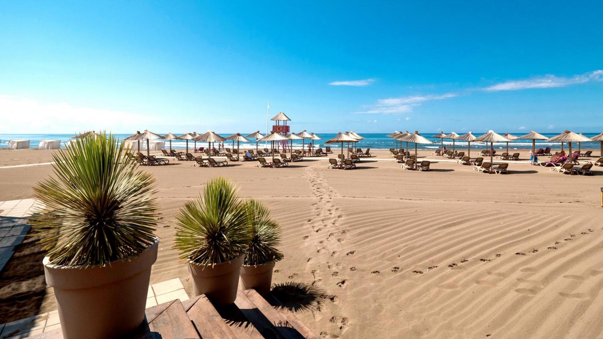 Azul Beach Resort Montenegro plašā smilšu pludmale ar salmu saulessargiem un zviļņiem Ulciņā — skats no terases uz Adrijas jūru