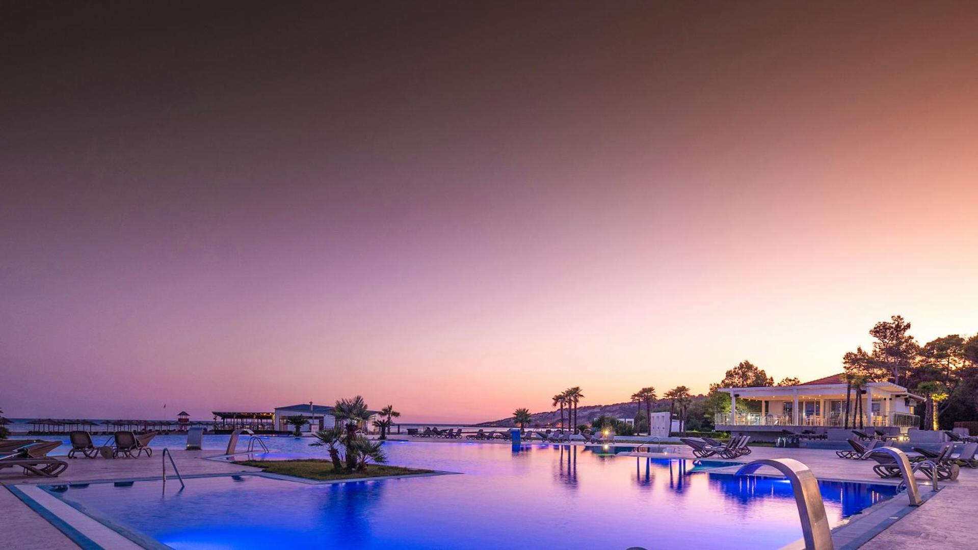 Azul Beach Resort Montenegro baseina zona saulrieta laikā ar izgaismotu ūdeni un purpursarkanām debesīm Melnkalnes piekrastē