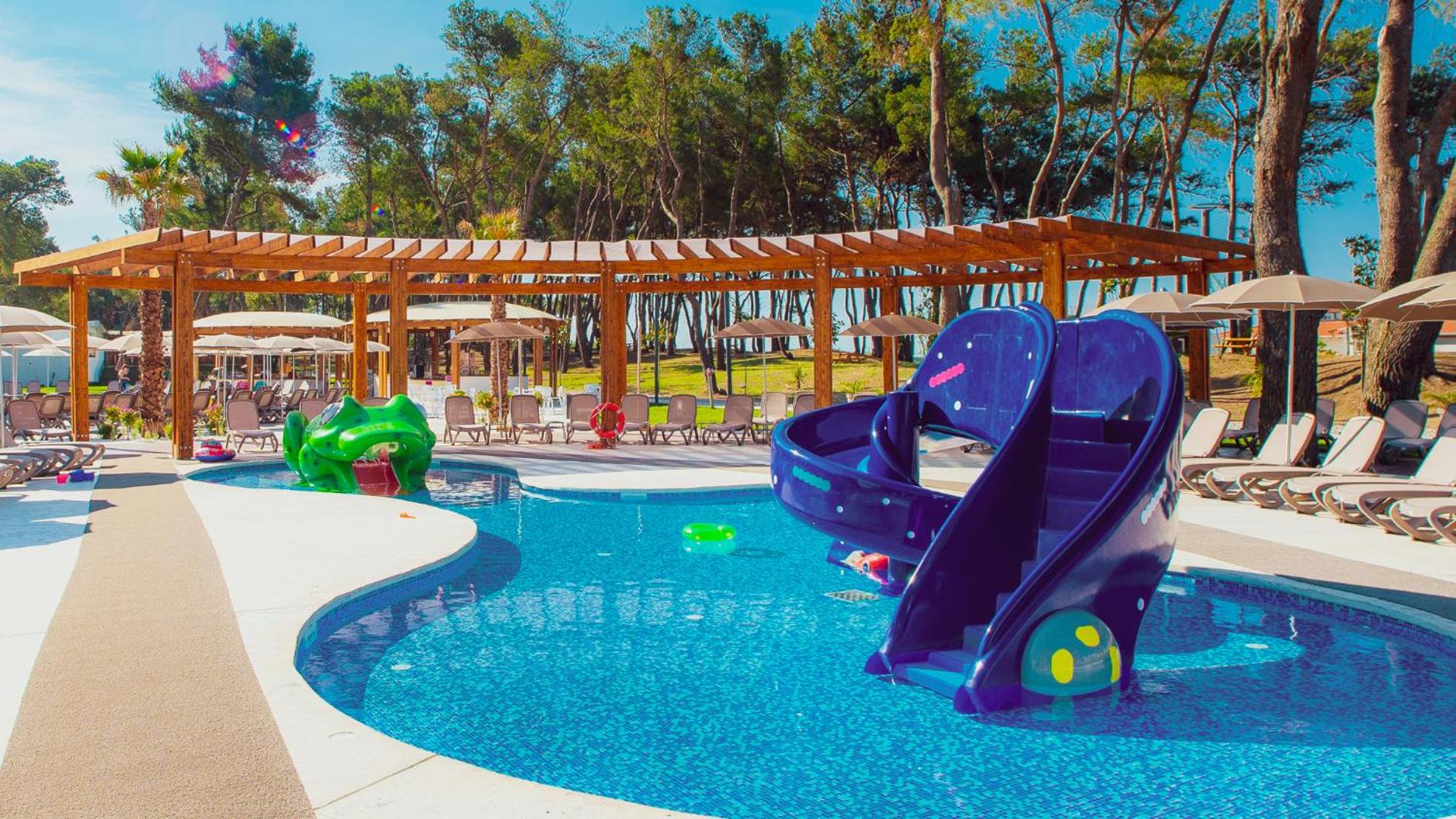 Azul Beach Resort Montenegro bērnu baseins ar krāsainiem slidkalniņiem un strūklakām, ko ieskauj sauļošanās krēsli un koka terase.