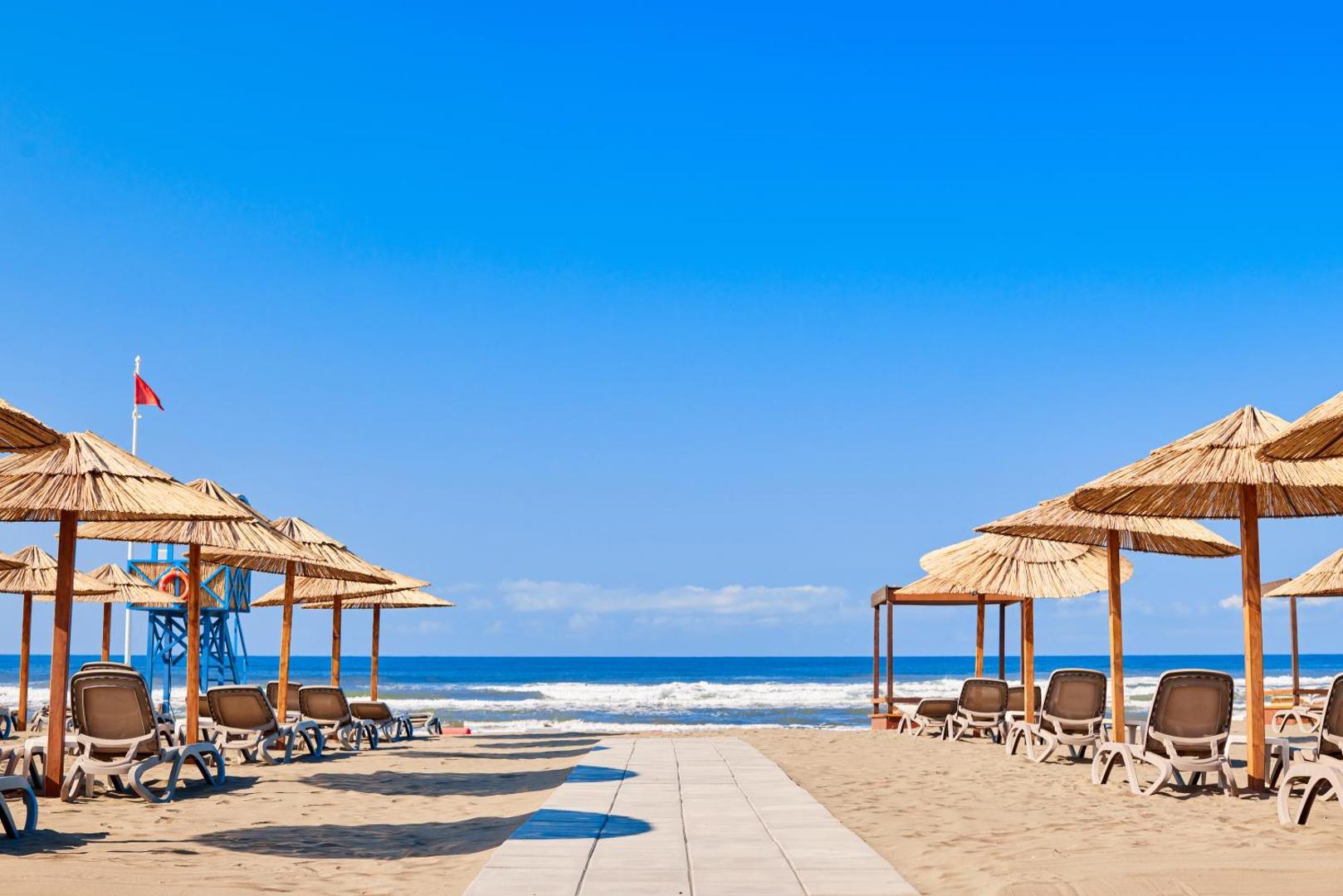 Azul Beach Resort Montenegro pludmales zona ar koka celiņu, salmu saulessargiem un glābēju torni zilās debess fonā