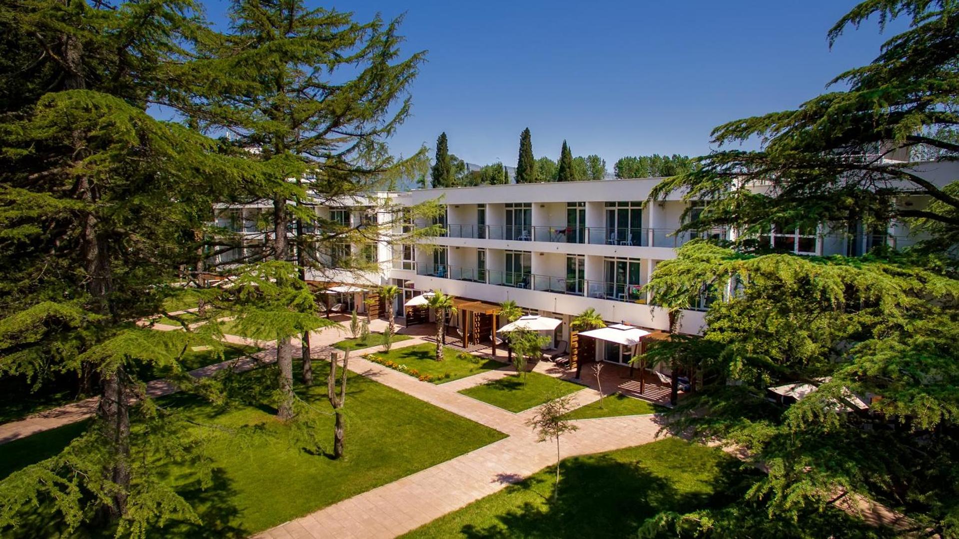 Azul Beach Resort Montenegro viesnīcas korpuss ar balkoniem, ko ieskauj kupli, zaļi koki un labiekārtoti zālieni
