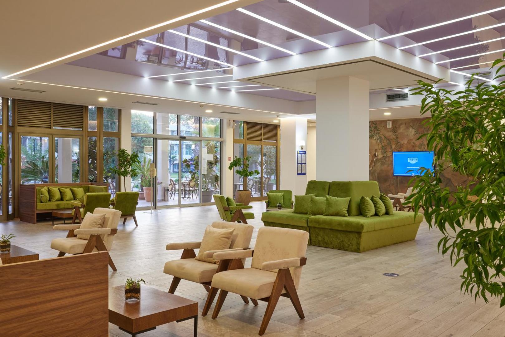 Azul Beach Resort Montenegro gaišais lobby bārs ar zaļiem dīvāniem, atpūtas krēsliem un lieliem panorāmas logiem uz dārzu