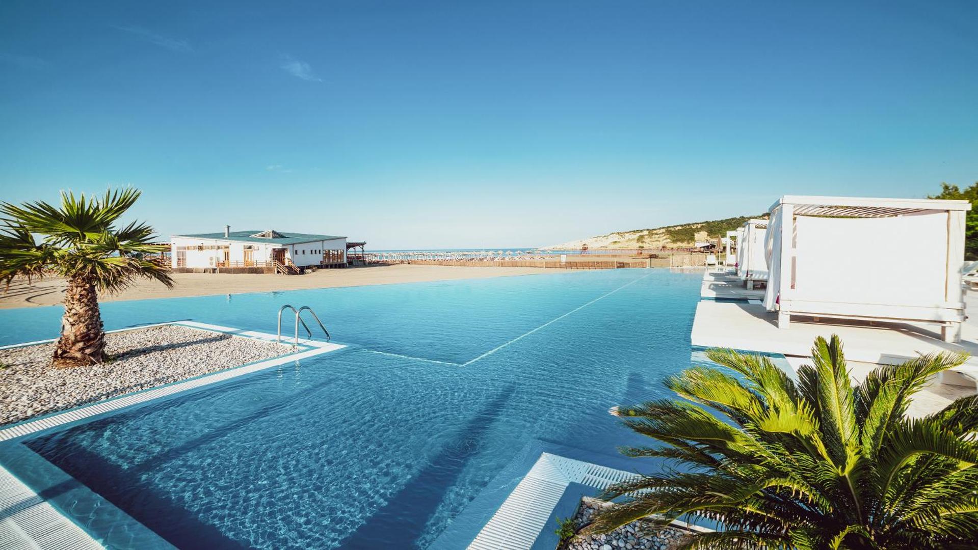 Azul Beach Resort Montenegro bezgalības baseins pie pludmales ar baltām atpūtas nojumēm un palmām Ulciņas piekrastē.