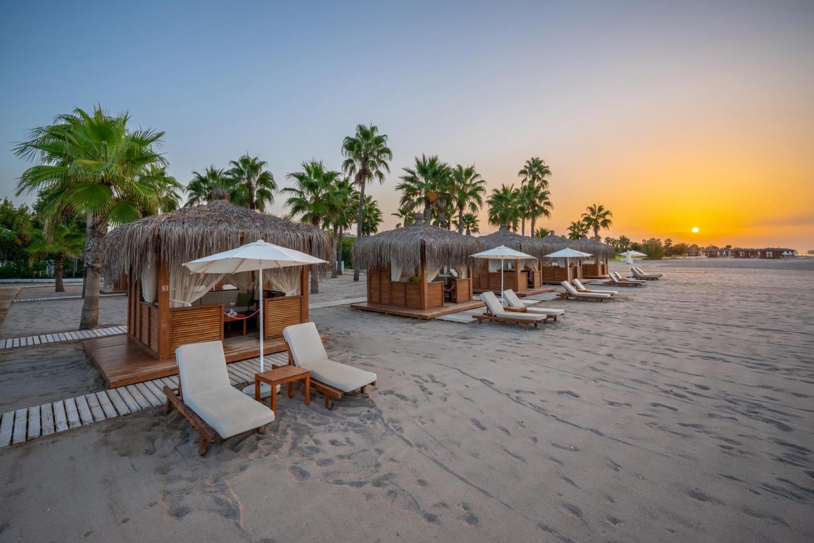 Selectum Family Resort Belek privātā pludmale saulrieta laikā ar koka bungalo, saulessargiem un ērtiem zviļņiem uz smiltīm