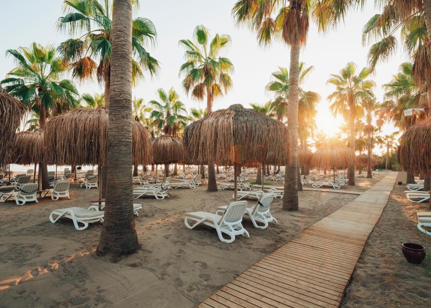 Selectum Family Resort Belek pludmales zona ar salmu saulessargiem, baltiem zviļņiem un palmām saulrieta gaismā Turcijas piekrastē