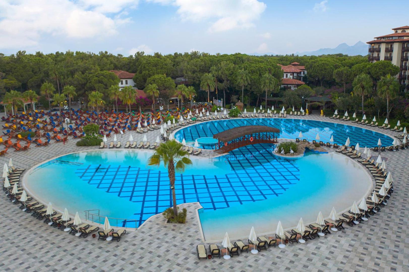 Selectum Family Resort Belek lielais āra baseins ar tiltiņu, saulessargiem un priežu mežu fonā Belekas reģionā, Turcijā.
