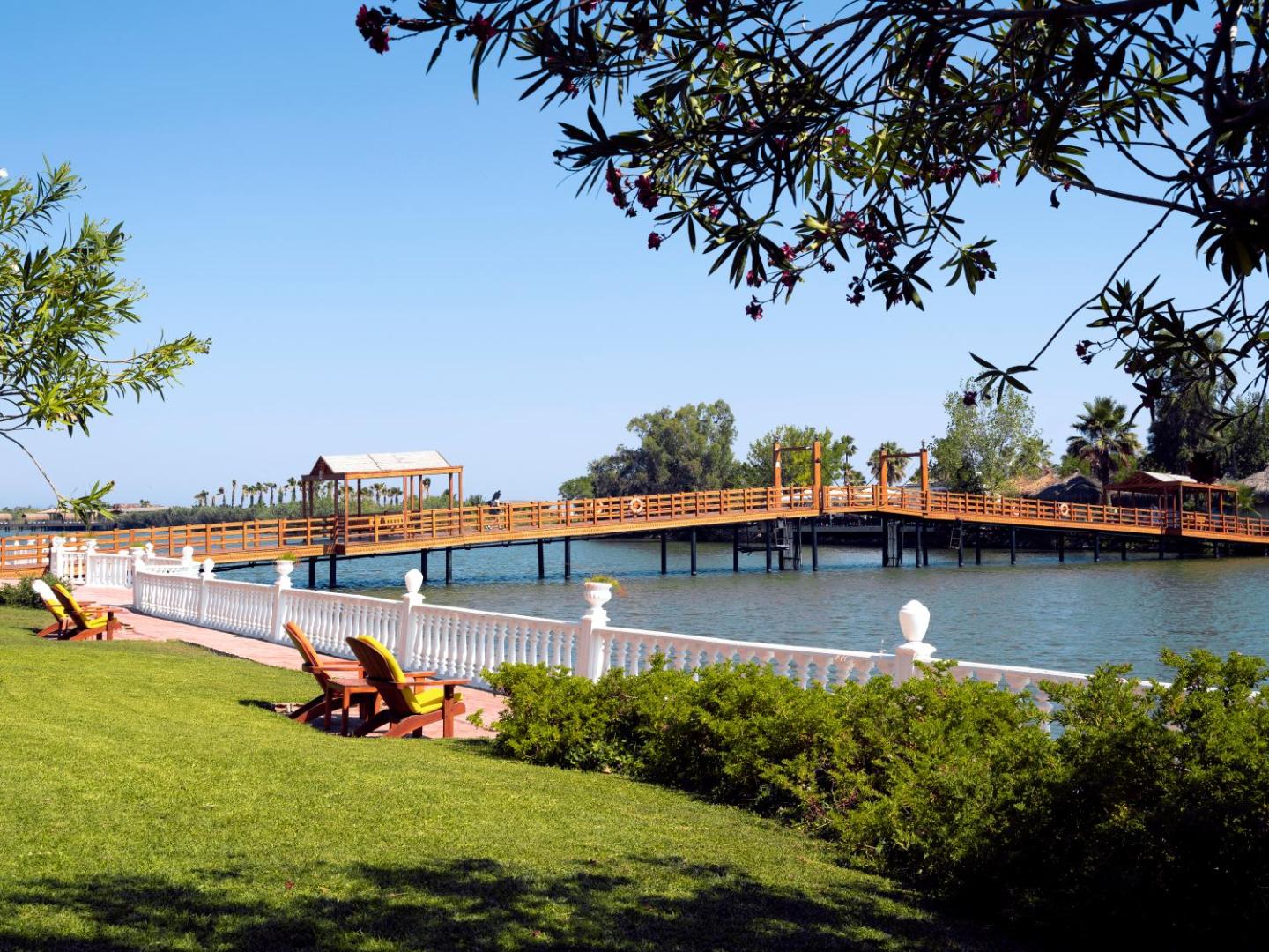 Koka tilts pār upi Selectum Family Resort Belek teritorijā — atpūtas krēsli zālienā pie ūdens malas