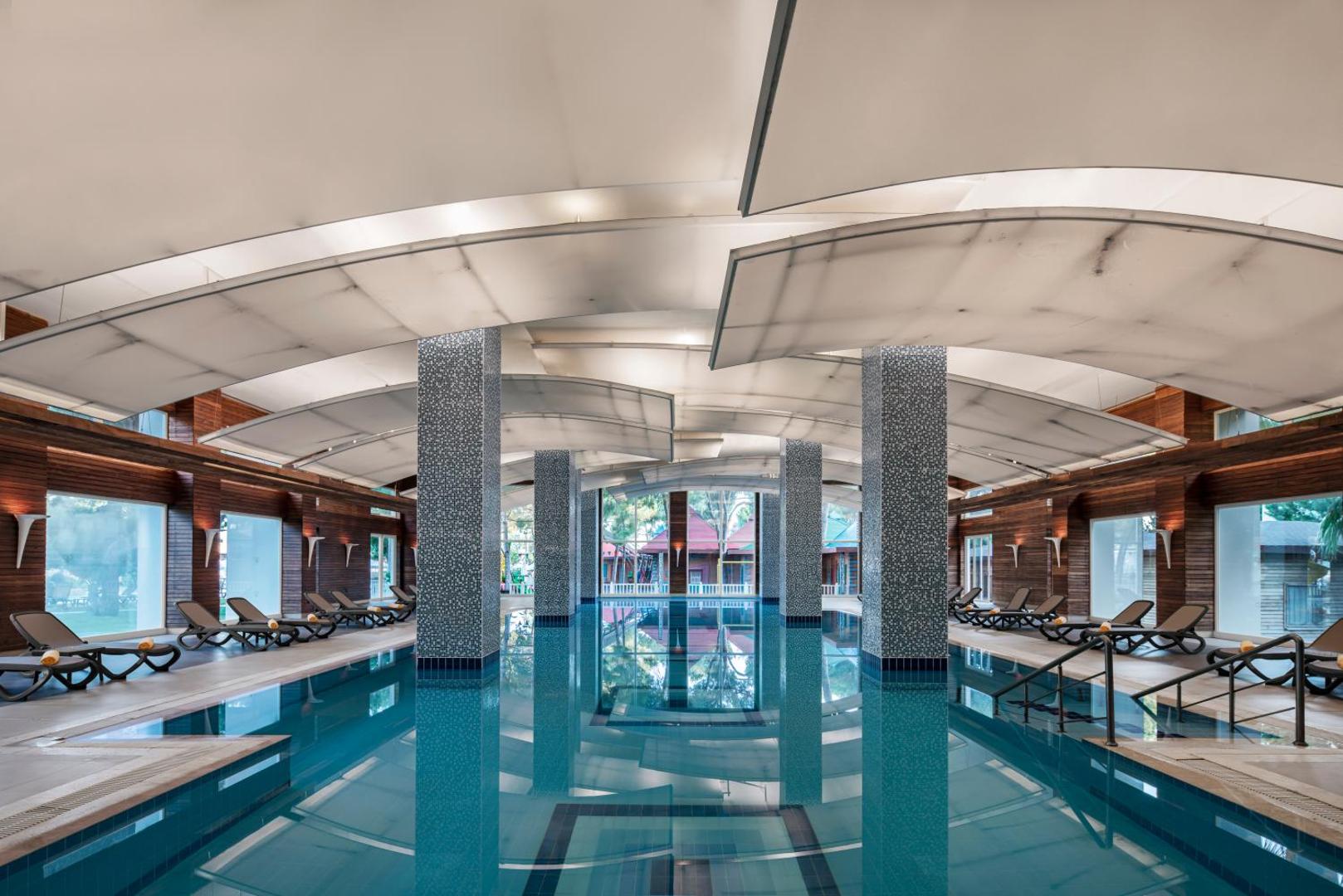 Papillon Ayscha iekštelpu baseins ar modernu griestu dizainu, mozaīkas kolonnām un atpūtas krēsliem relaksācijai spa zonā