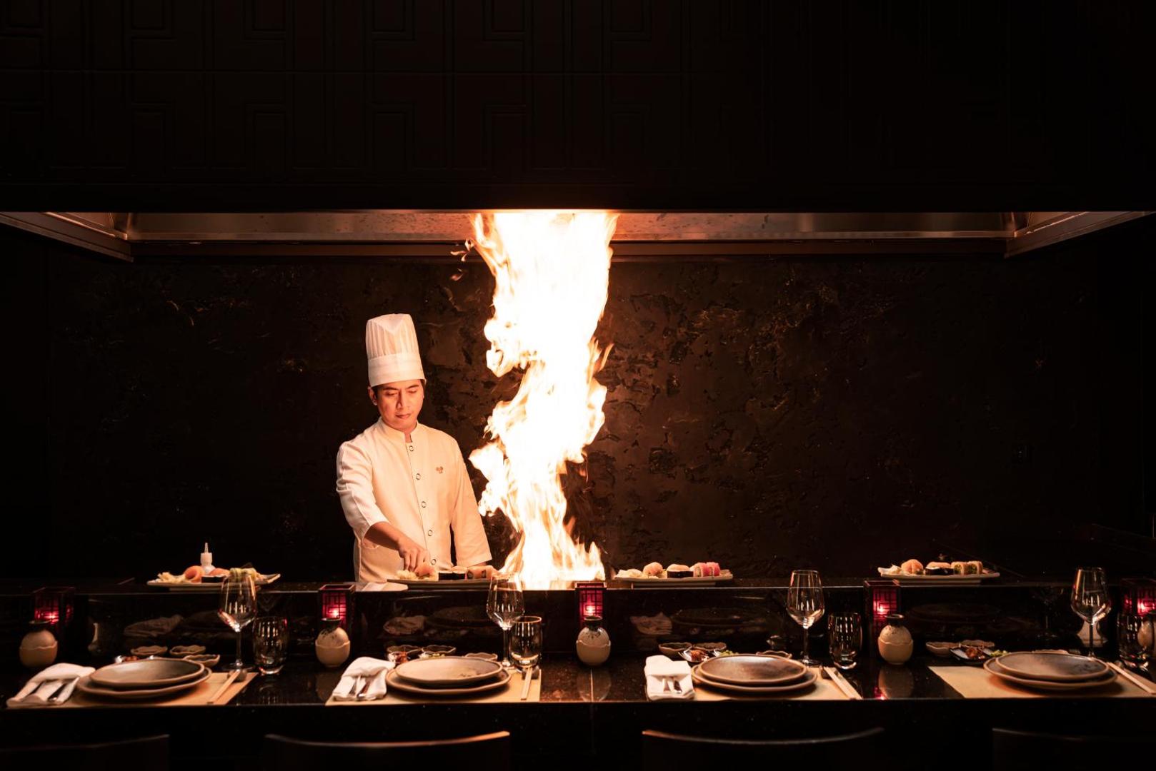 Šefpavārs gatavo ēdienu uz atklātas uguns Papillon Ayscha Teppanyaki restorānā, radot iespaidīgu liesmu šovu viesu priekšā