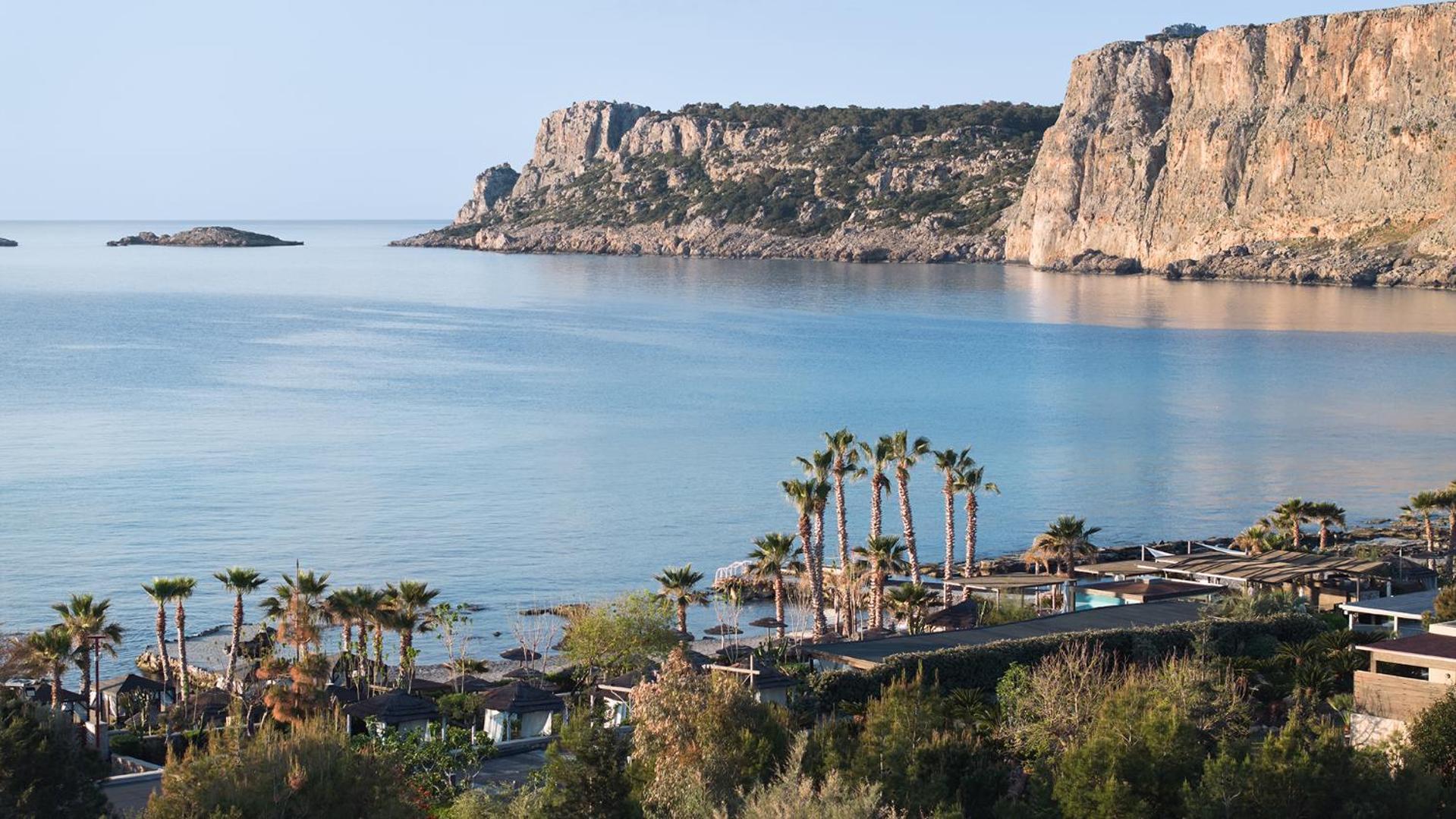 Panorāmas skats no Aquagrand Exclusive Deluxe Resort uz Navarones līci Rodas salā — piekrastes klintis, palmas un dzidrs jūras ūdens