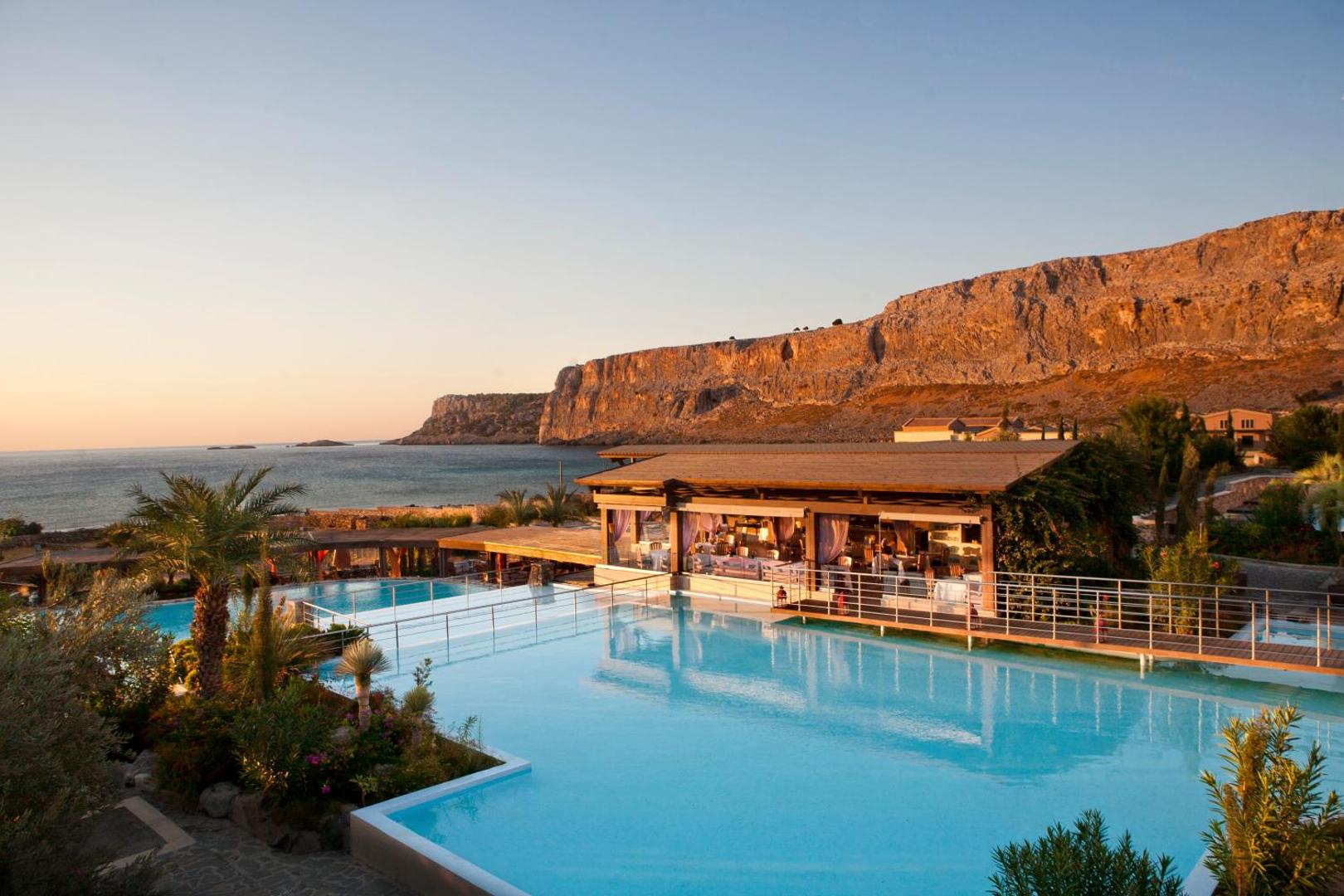 Aquagrand Exclusive Deluxe Resort āra baseins saulrietā ar skatu uz klintīm un jūru Lindosā — restorāna zona pie ūdens