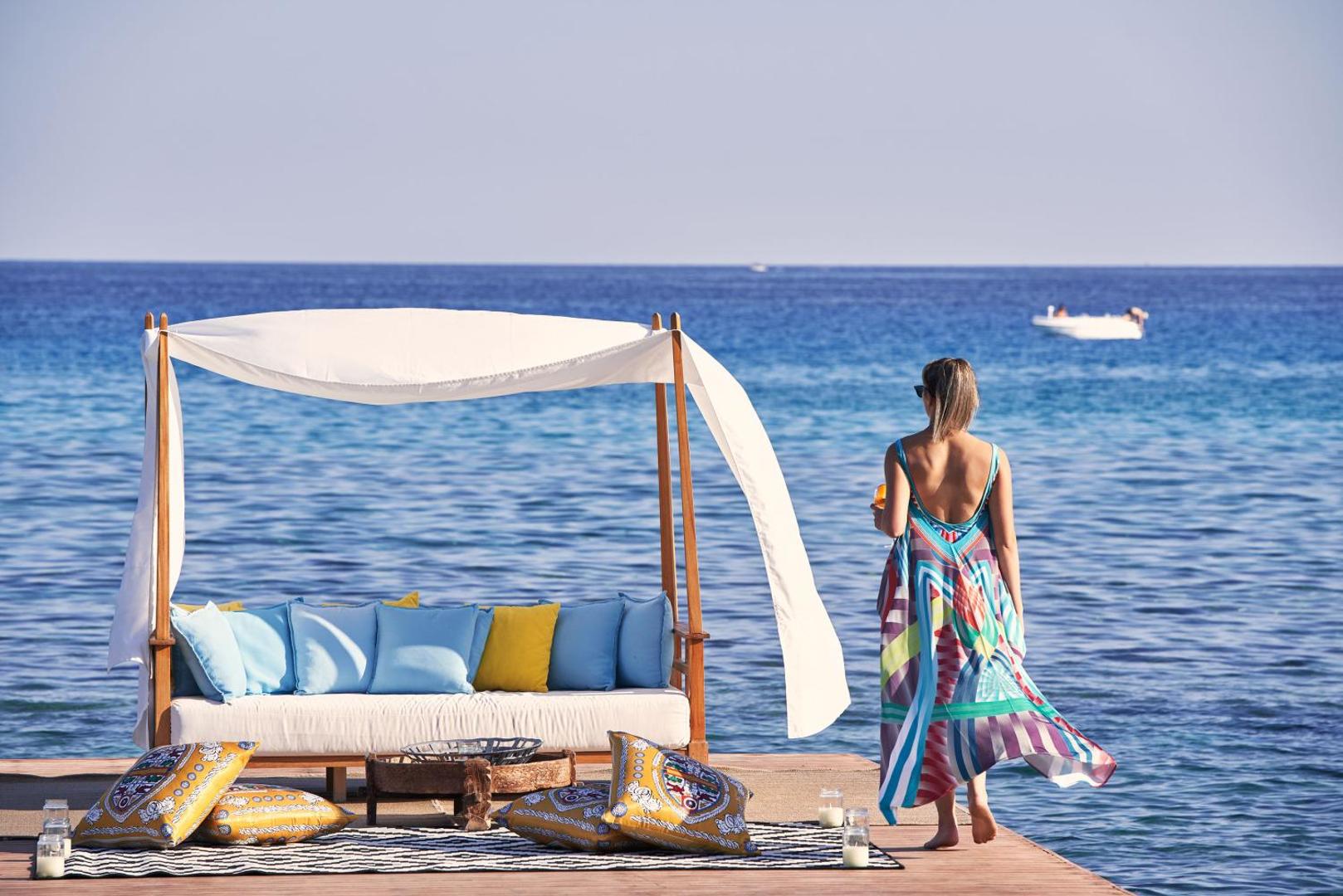 Aquagrand Exclusive Deluxe Resort atpūtas zona uz koka platformas pie jūras Rodas salā ar dīvānu un krāsainiem spilveniem