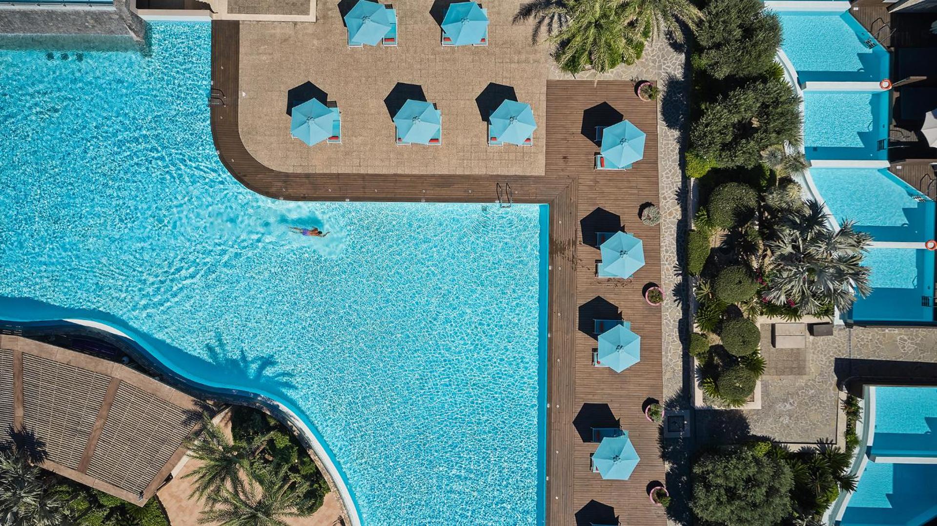 Aquagrand Exclusive Deluxe Resort baseina zonas skats no augšas — tirkīza ūdens, koka klājs ar saulessargiem un atpūtas zonu