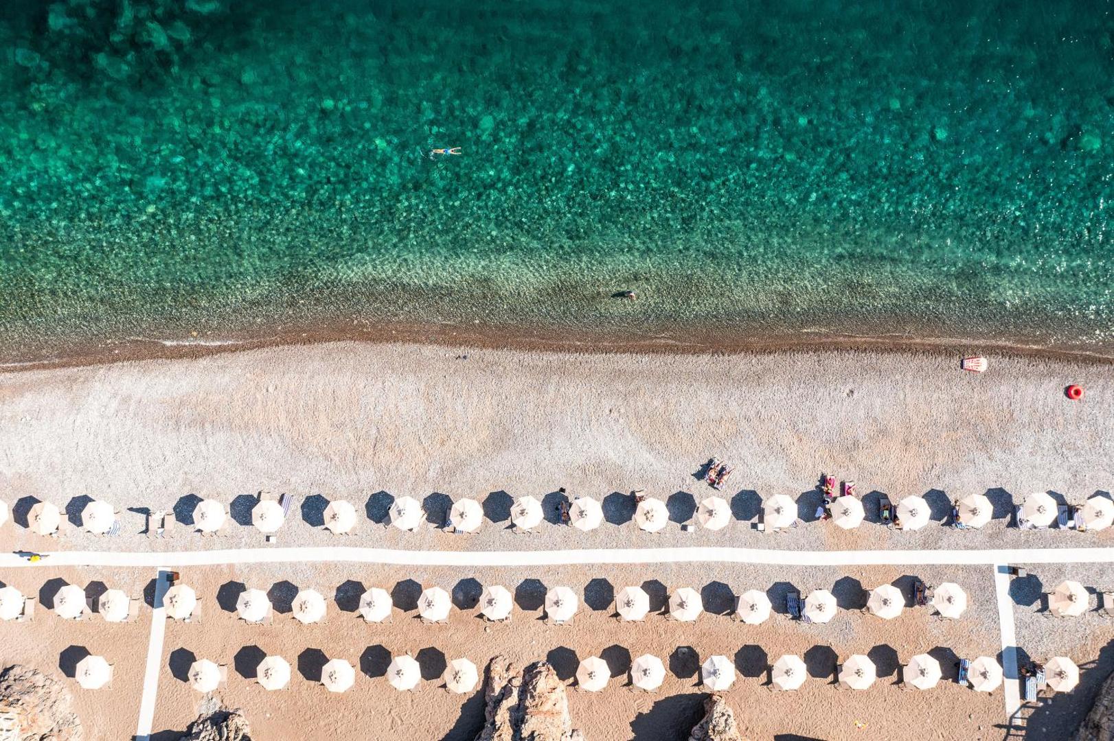 The Chedi Lustica Bay privātā oļu pludmale no putna lidojuma — tirkīza ūdens, balti saulessargi un sakopta krasta līnija