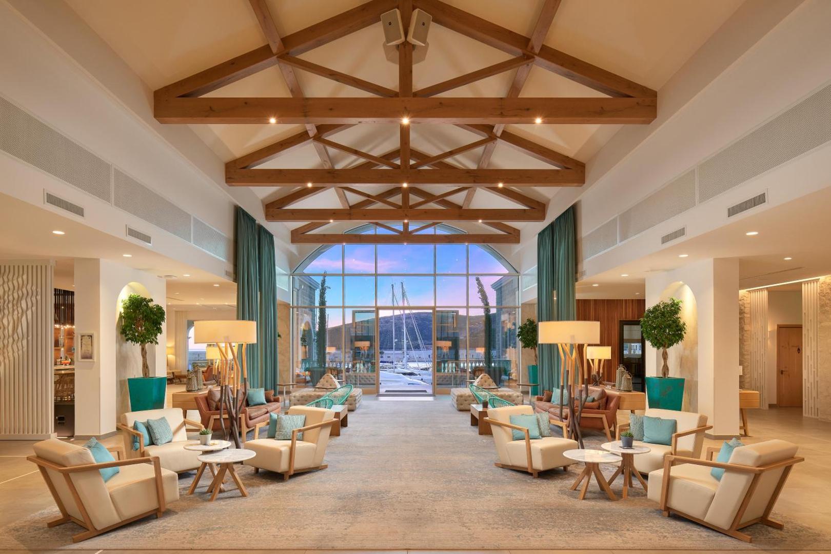 The Chedi Lustica Bay viesnīcas plašais vestibils ar augstiem koka griestiem un panorāmas logu uz jahtu piestātni Melnkalnē