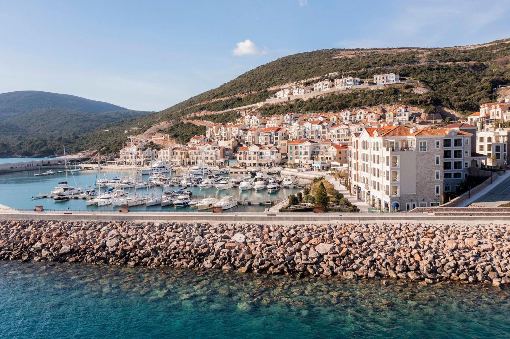 The Chedi Lustica Bay viesnīcas galvenā ēka un jahtu osta saulrietā — akmeņains mols priekšplānā un kalnu ainava fonā