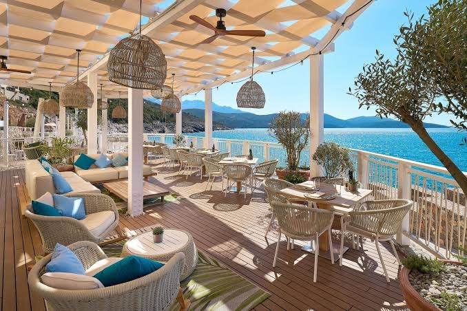 The Chedi Lustica Bay plašā restorāna terase ar koka klāju, pītiem krēsliem un nojumi piekrastē ar skatu uz kalniem