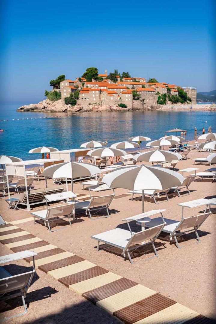 California by Aycon pludmale ar gaišiem saulessargiem un zviļņiem uz smilšu un oļu krasta Sveti Stefan kūrortā