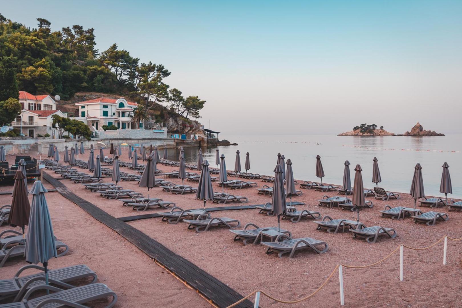 Hotel Ami Budva Petrovac privātā pludmale ar sakārtotām sauļošanās krēslu rindām, saulessargiem un skatu uz klinšainām salām jūrā.