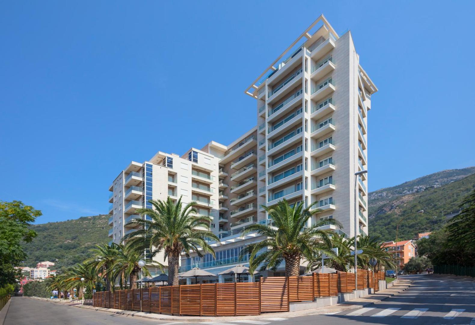 Hotel Ami Budva Petrovac modernā ēka Melnkalnē ar palmām un balkoniem, skats no ielas puses saulainā dienā