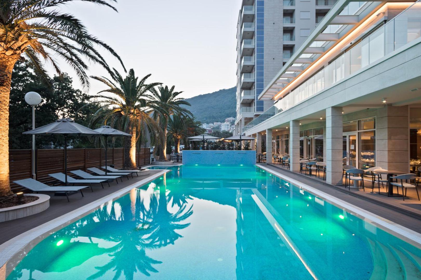 Hotel Ami Budva Petrovac āra baseins vakarā ar apgaismojumu, palmām un viesnīcas fasādi Petrovacas piekrastē