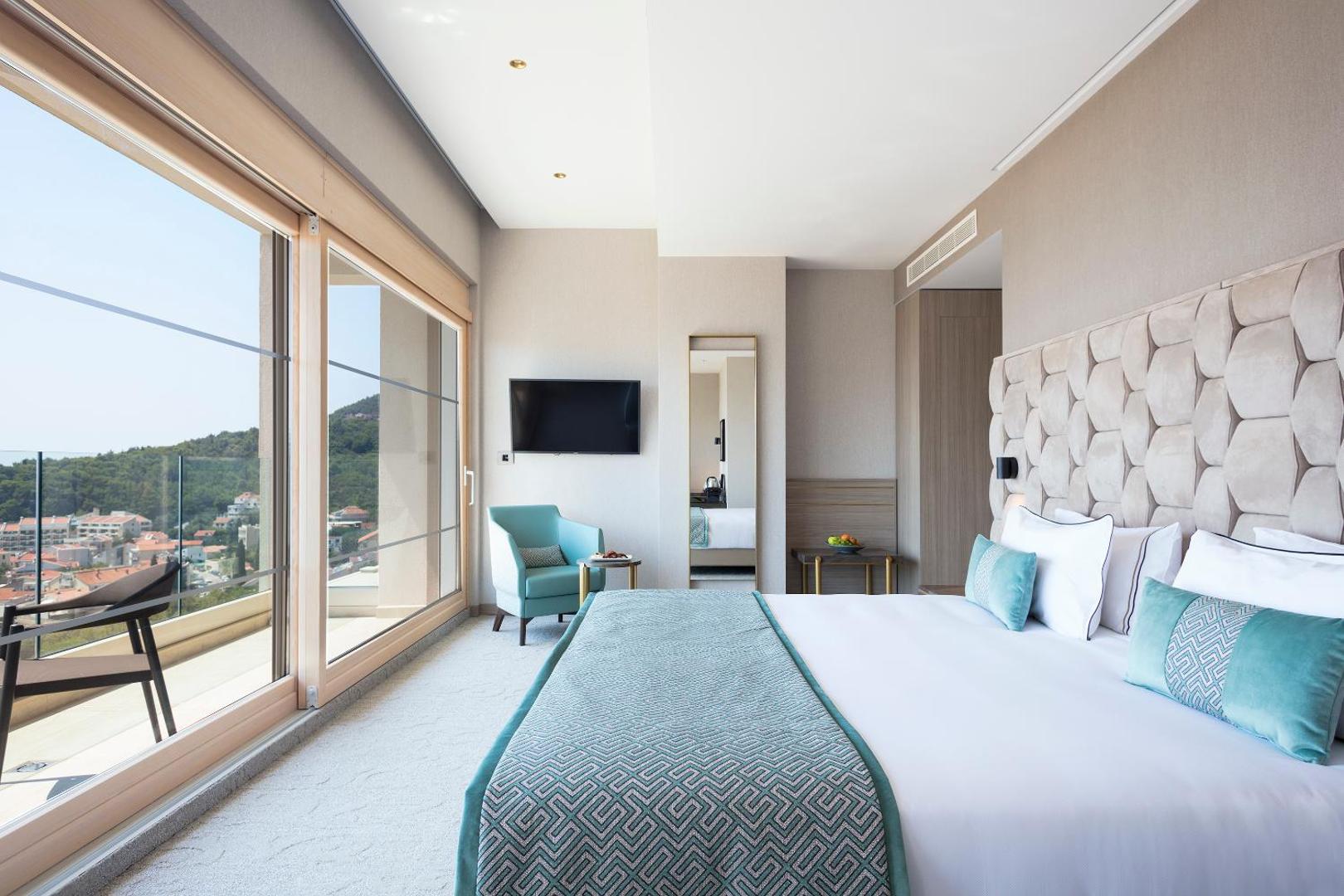 Mūsdienīgs Hotel Ami Budva Petrovac numura interjers ar mīkstu galvgali, tirkīza akcentiem un plašu balkonu ar skatu uz pilsētu.