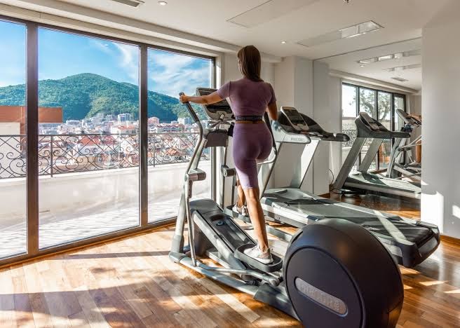 Viesnīcas Iberostar Waves Slavija sporta zāle ar skrejceļiem un eliptiskajiem trenažieriem pie lieliem logiem ar skatu uz Budvu