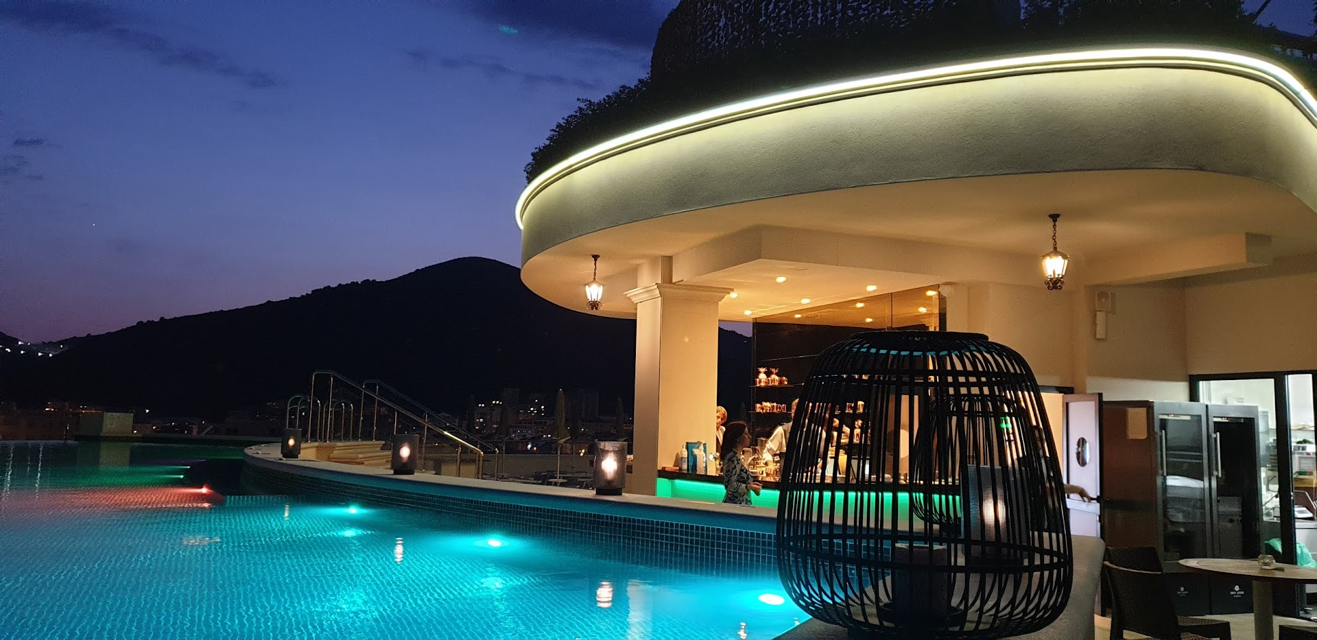 Iberostar Waves Slavija jumta bārs un baseins vakarā ar izgaismotu interjeru, modernām lampām un skatu uz kalniem krēslā