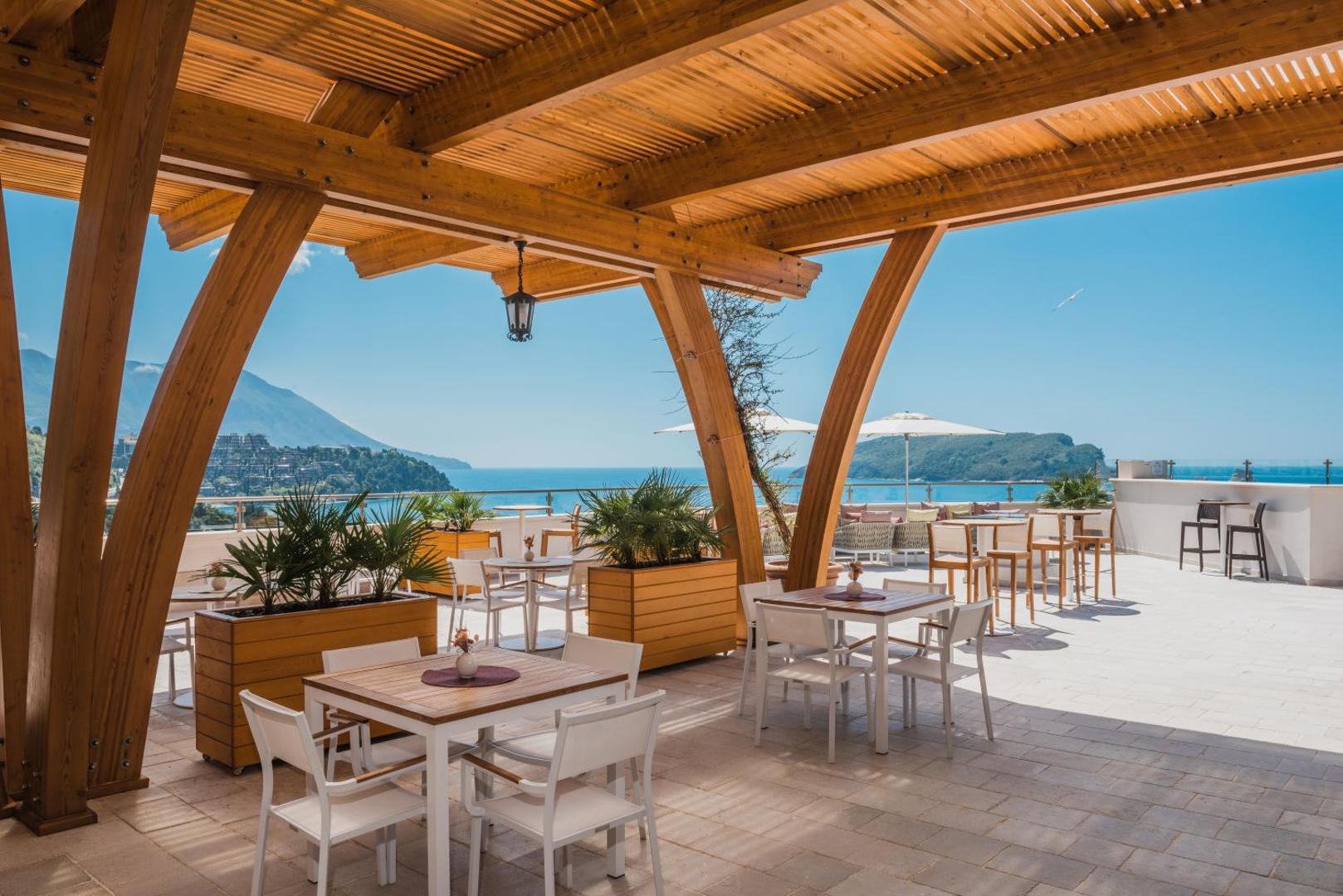 Iberostar Waves Slavija viesnīcas āra terase ar koka konstrukcijām, galdiņiem un skatu uz jūru un Budvas piekrastes kalniem