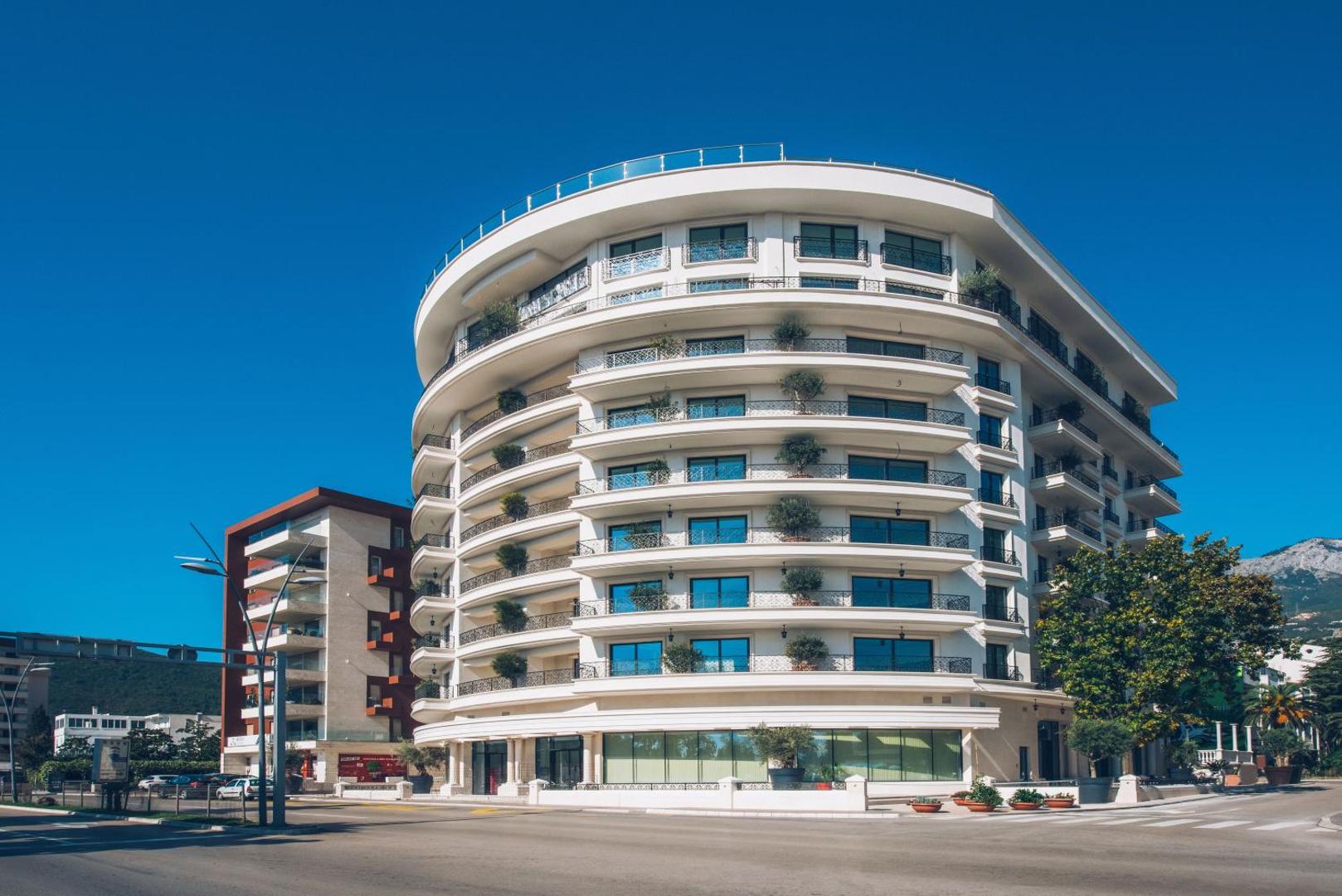 Iberostar Waves Slavija viesnīcas modernā fasāde Budvā, Melnkalnē — balta, pusapaļa ēka ar balkoniem un skatu uz pilsētu un kalniem