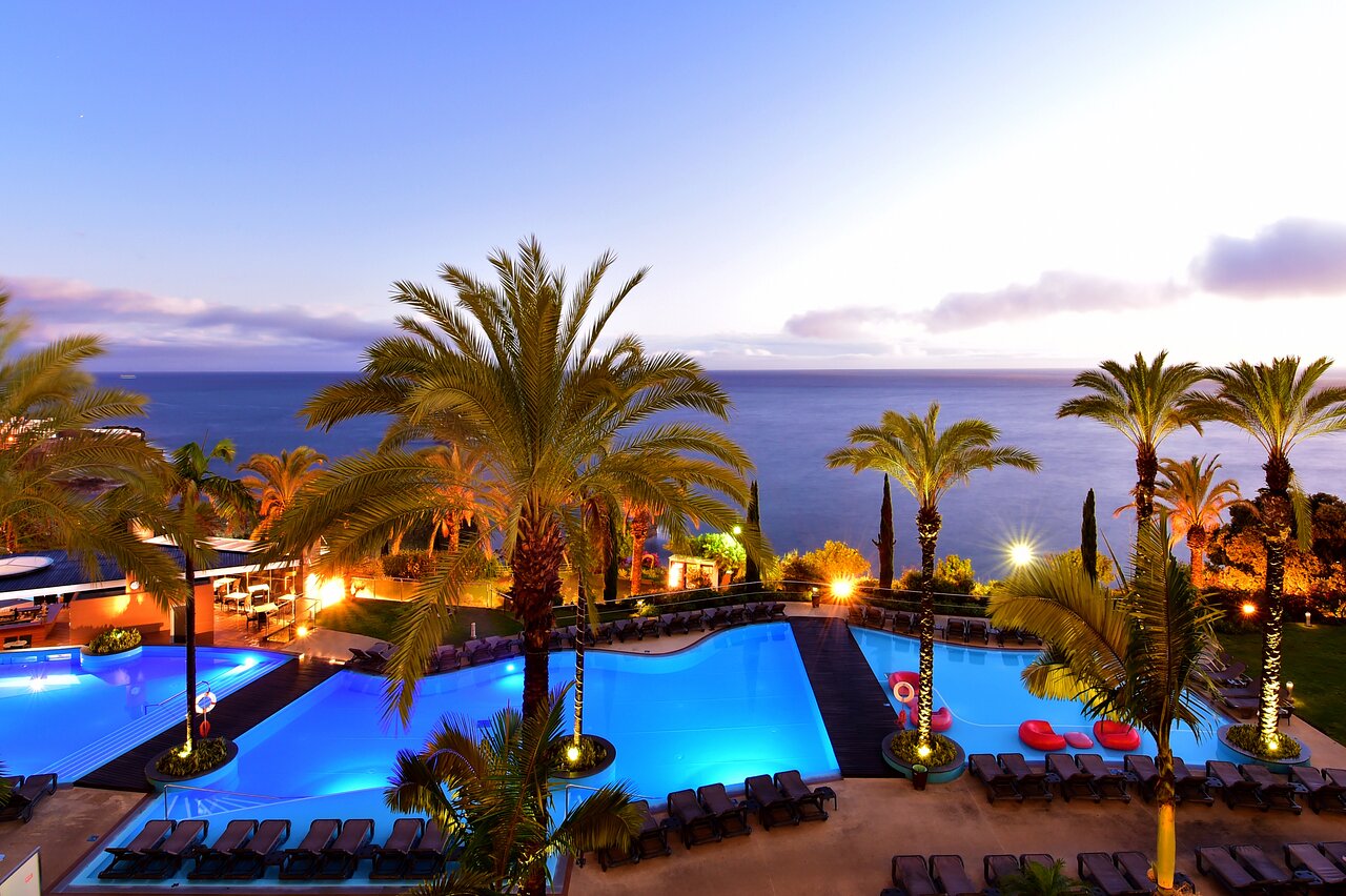 Peldbaseins ar zilu ūdeni un palmu kokiem pie jūras, apgaismots vakara gaismā. Ideāla vieta relaksācijai un atpūtai. panoramic view with beach and pools