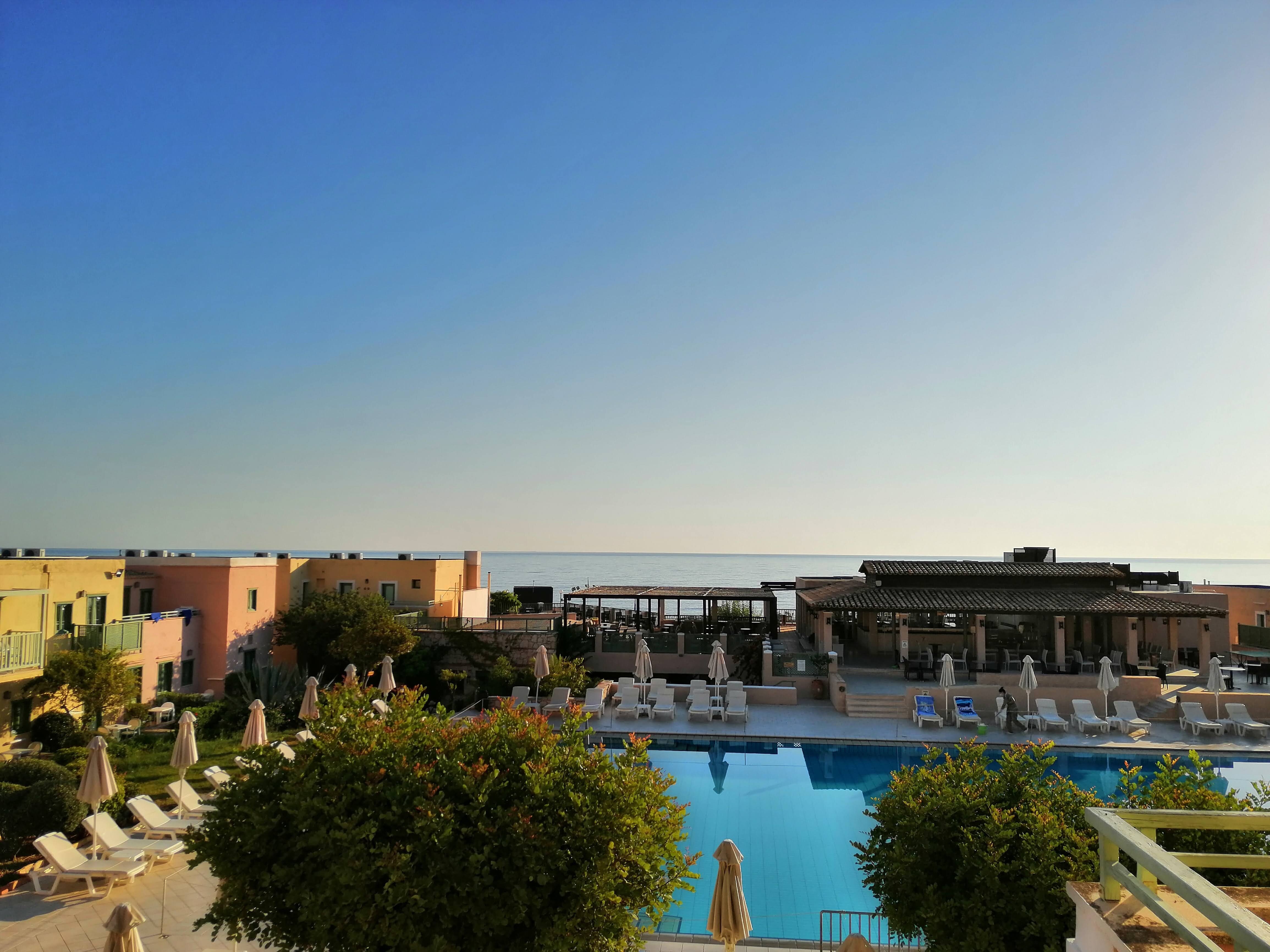 Jauka pludmales viesnīca ar baseinu un skatu uz jūru, apkārt ir sauļošanās krēsli un zaļumi. Skats ir mierīgs ar skaidru debesu zilo krāsu. panoramic view with beach and pools