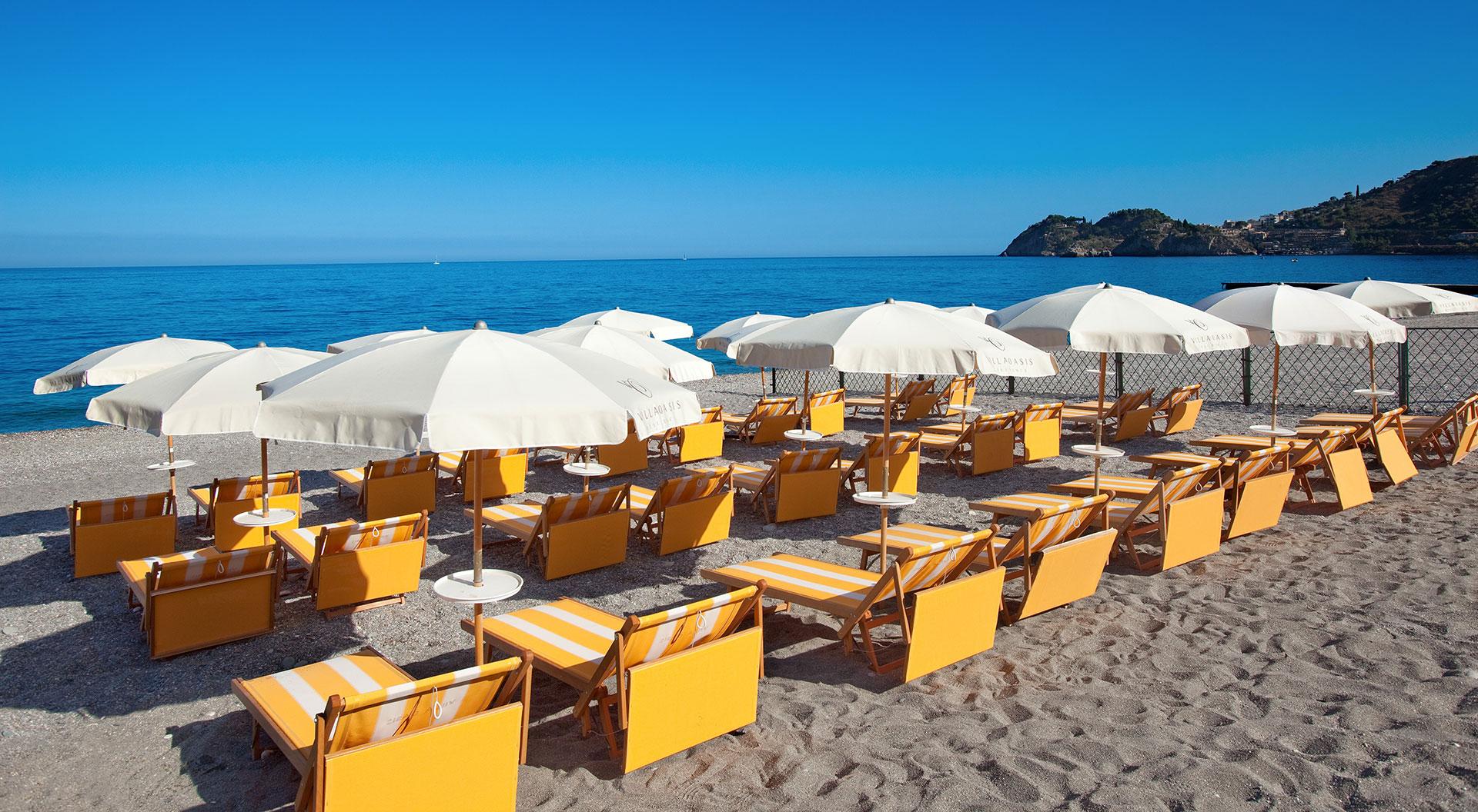 Pludmales atpūtas zona ar dzeltenām sauļošanās krēslām un baltām saules omītēm, kas izkārtotas pie jūras. Skats uz mierīgu ūdeni un skaidru zilu debesu fonu.
