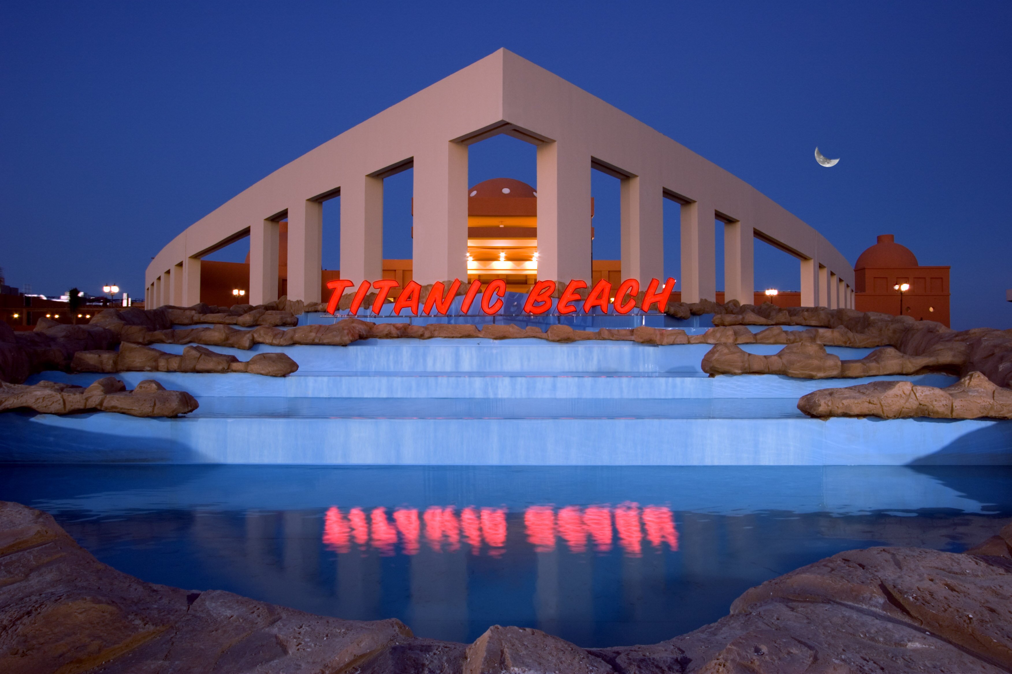 TITANIC BEACH viesnīcas priekšpuse ar krāsainu zīmolu un baseinu, apgaismota nakts laikā. panoramic view with beach and pools