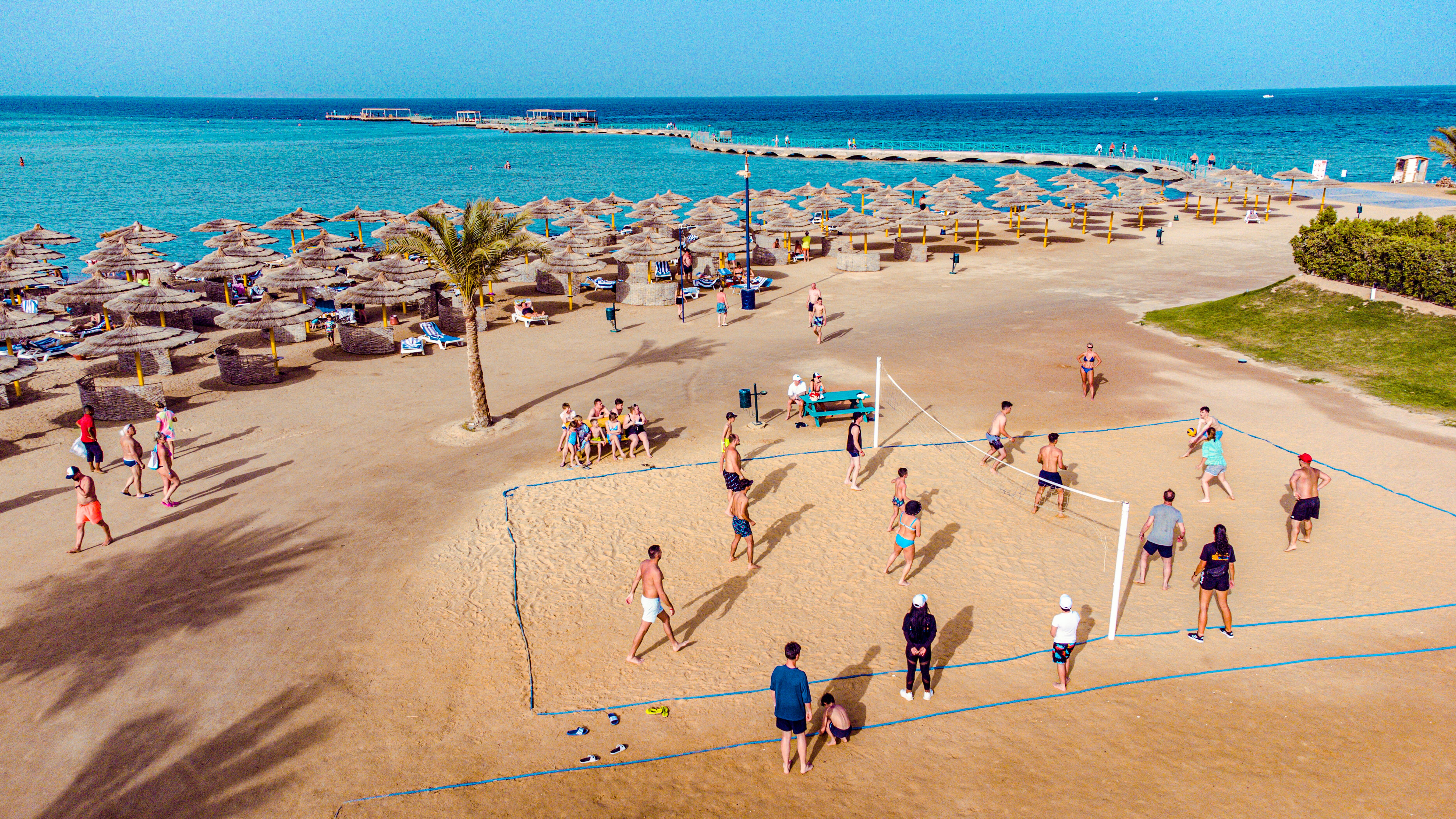 Pludmales volejbola spēlētāji aktīvi izbauda spēli saulainā pludmalē, apkārt citi apmeklētāji relaksējas zem saulessargiem. panoramic view with beach and pools