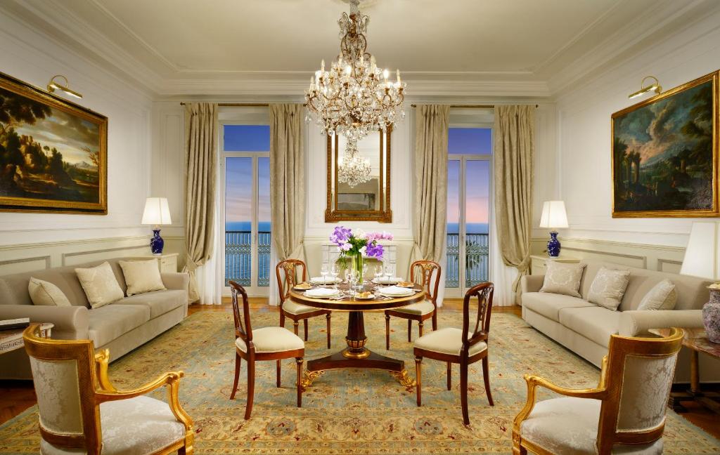 Elegants viesistabas interjers ar jūru redzamu caur logu, dekorēts ar gleznām un greznu kristāla lustru. Centrālais galds ir klāts ar ziediem un galda piederumiem, radot mājīgu atmosfēru.