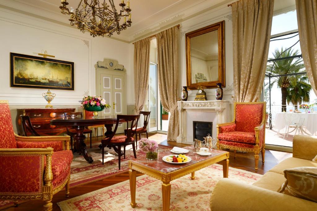 Elegantā viesistaba ar greznām mēbelēm un lieliem logiem, caur kuriem redzama daba. Interjeru rotā mākslas darbi un grezni dekoratīvie elementi.