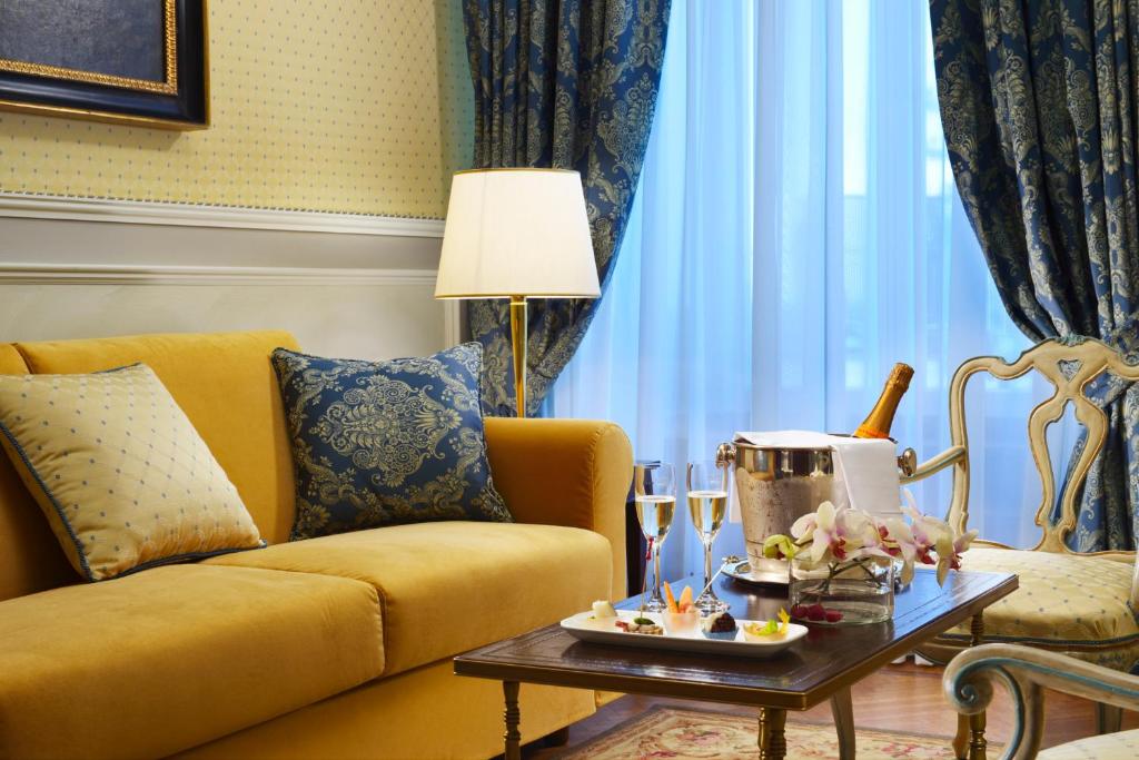 Eleganta viesnīcas viesistaba ar dzeltenu dīvānu, zilu aizkaru un galdu, kurā ir šampanietis un delikāti ēdieni. Interjers izceļas ar greznu un mājīgu atmosfēru.