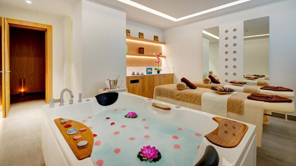 Mūsdienīgs spa telpa ar džakuzi, kas piepildīts ar ziedlapiņām un rozēm, blakus masāžas galdiem un relaksējošu apgaismojumu.
