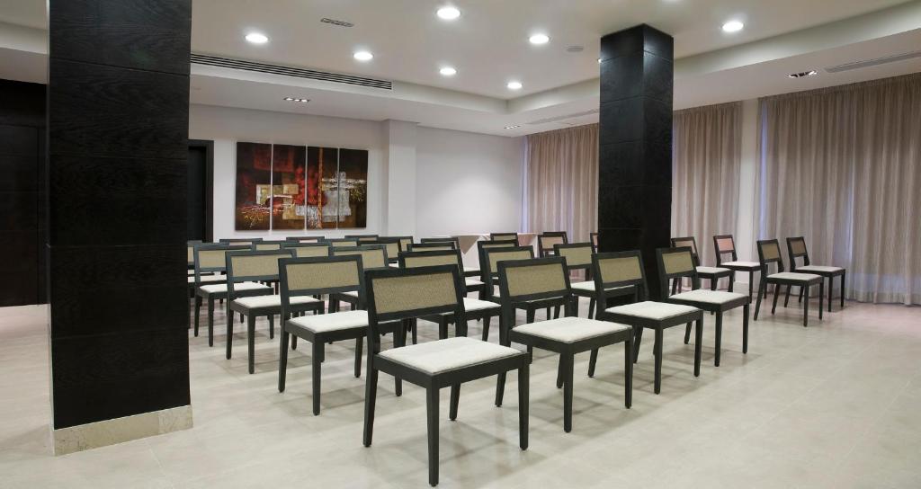 Konferenču telpa ar rindu krēsliem un gaismas apgaismojumu, kas rada modernu un profesionālu atmosfēru. Telpa ir aprīkota ar logiem un dekoratīvām gleznām uz sienām.